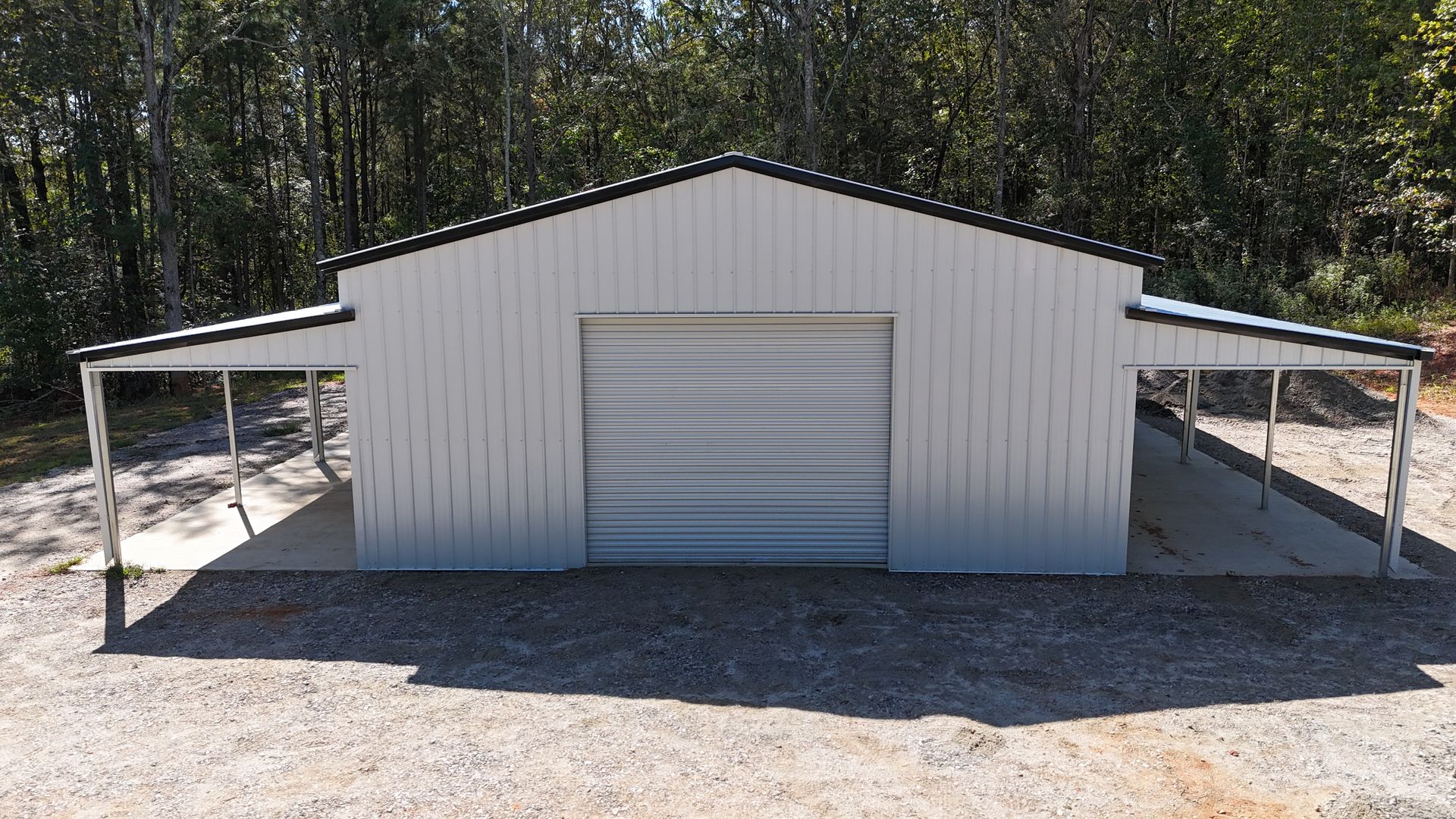 White metal shed with roll-up door, flanked by two carports on a gravel driveway, set against trees.