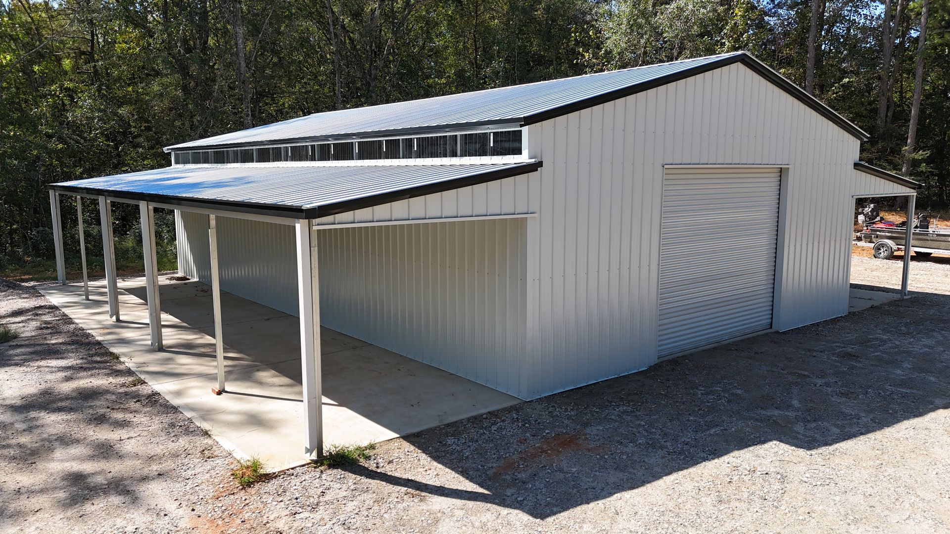 White metal barn with a covered overhang and concrete base, set in front of trees.