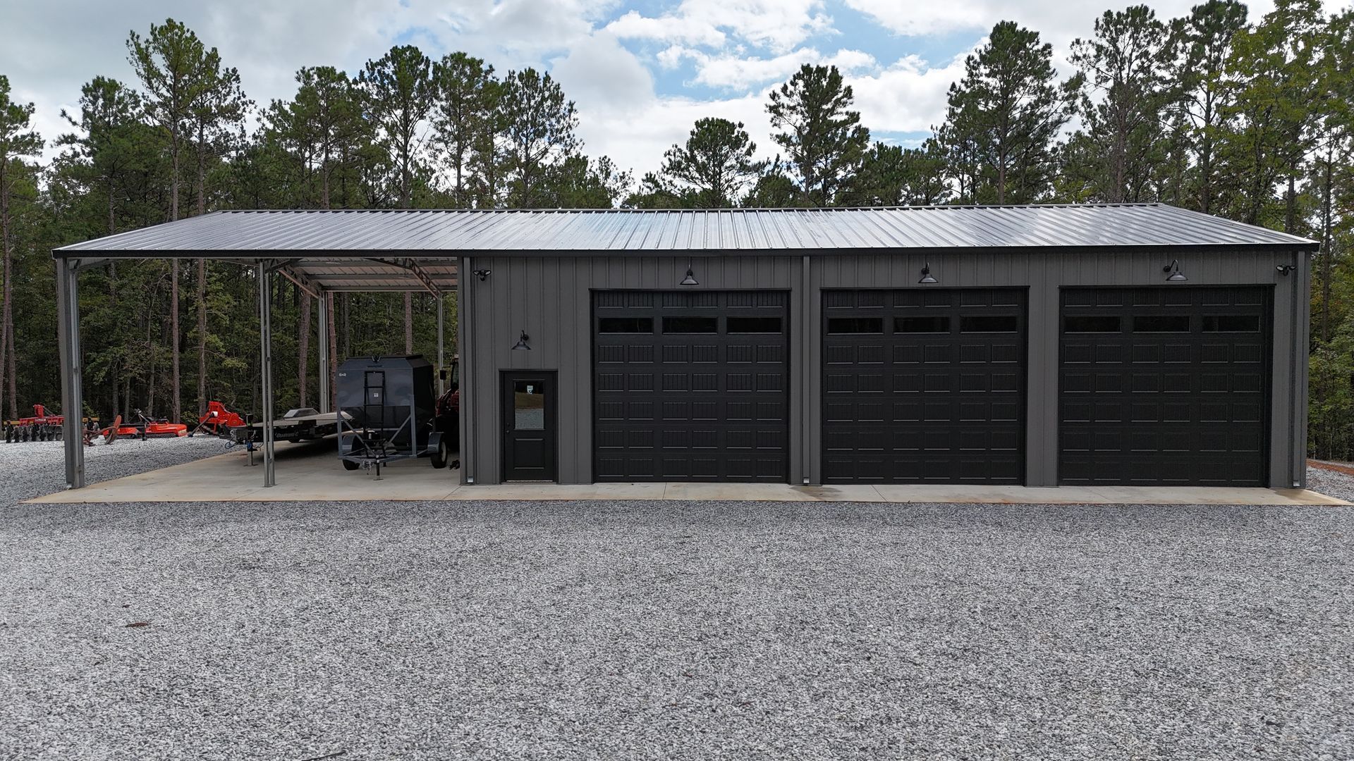 Gray metal building with three garage bays and a carport; set on gravel, trees in the background.