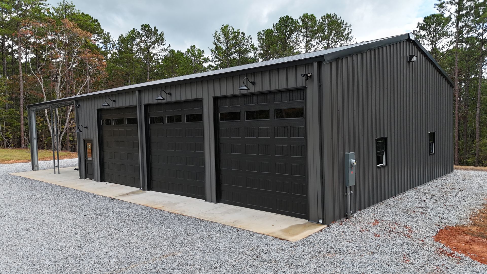 Dark gray metal garage with three bays and black garage doors. Gravel driveway, set in trees.
