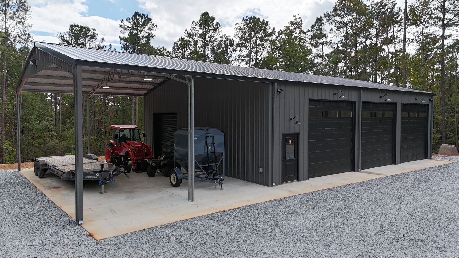 Gray metal workshop with carport, housing vehicles on a gravel lot; trees in background.