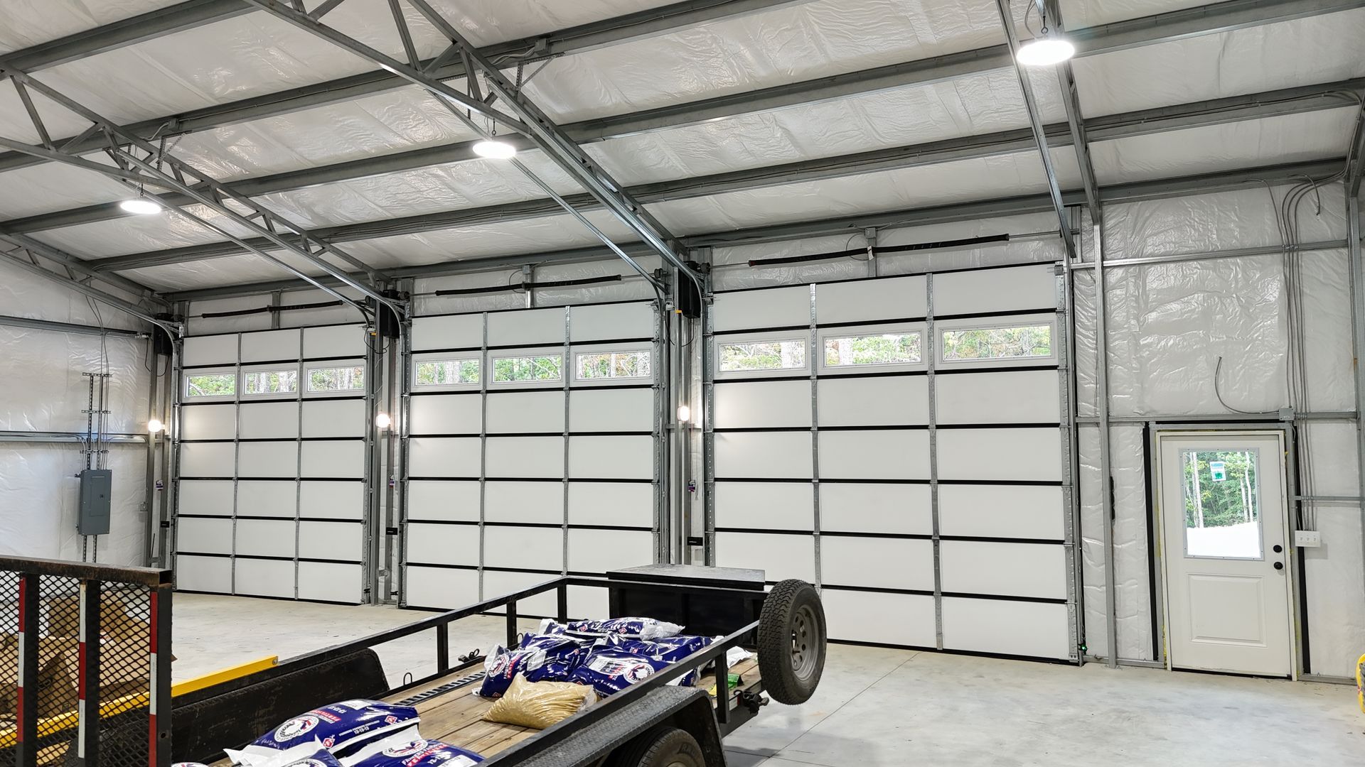 Three garage doors and a white door inside a metal-framed building; a trailer sits in the foreground.