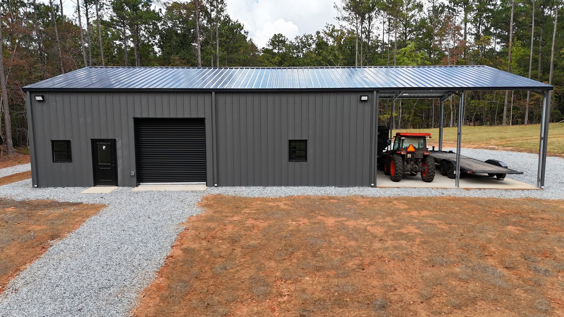 Gray metal building with garage door, covered side for tractor, gravel drive, trees.