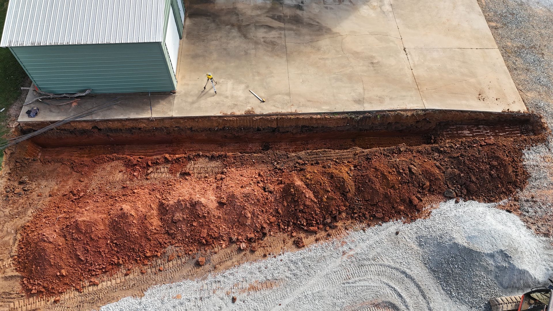 Trench dug in red soil next to concrete slab, small building in background, and pile of gravel.