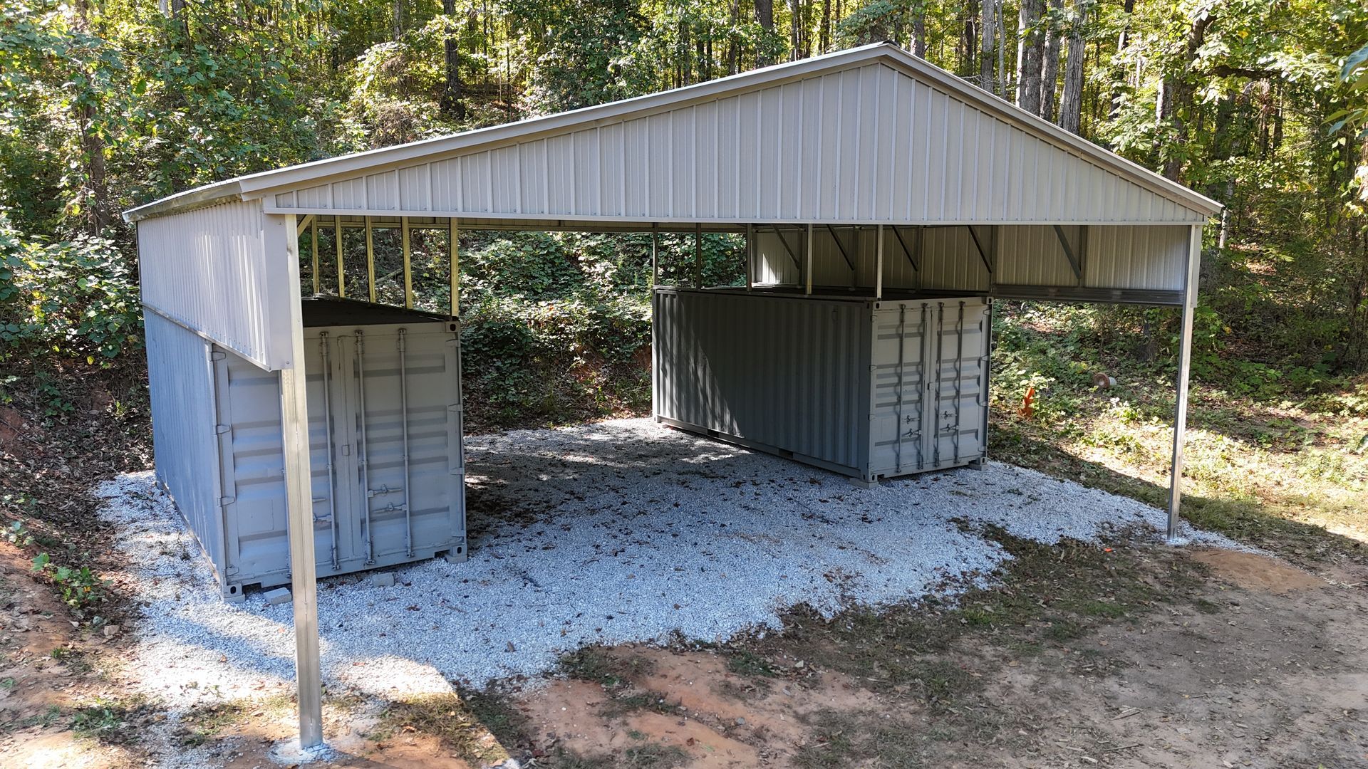 Metal carport sheltering two gray shipping containers on gravel. Trees in background.