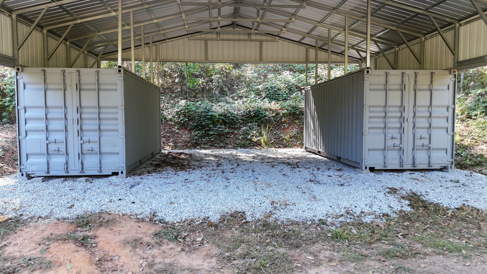 Two gray shipping containers under a metal roof shelter on a gravel bed.