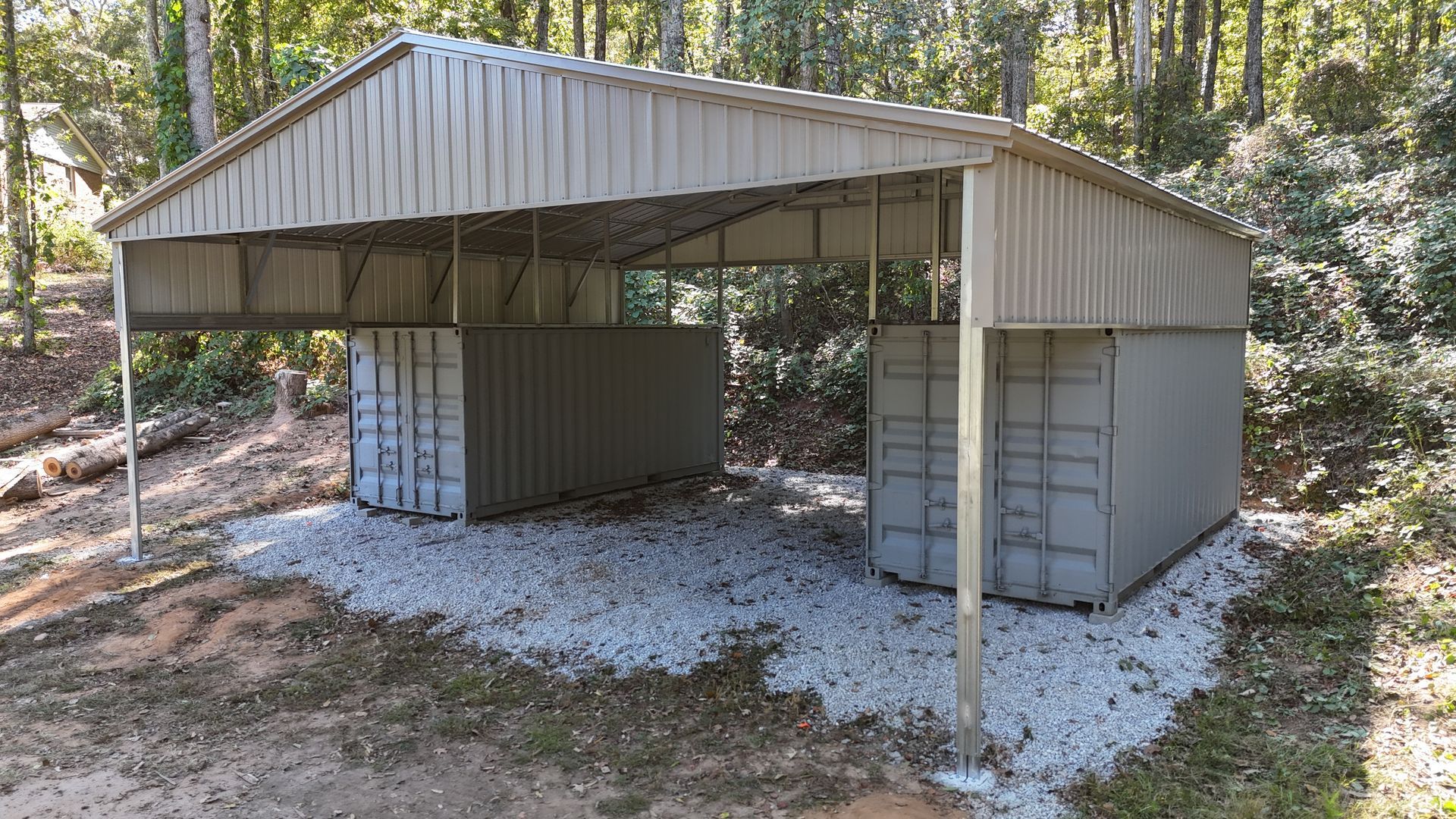 Metal carport with corrugated roof sheltering two shipping containers on a gravel base.
