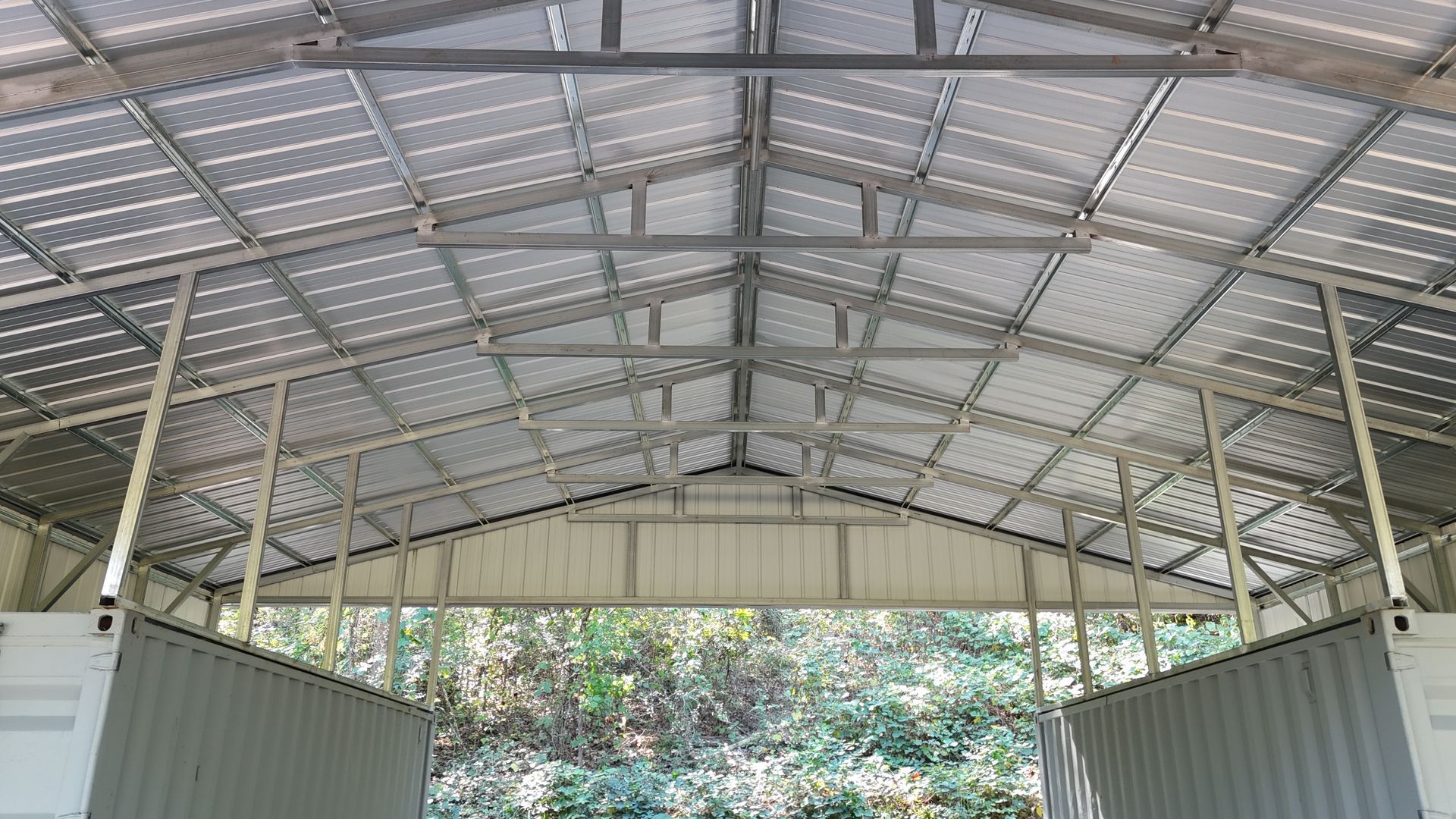 Steel-framed structure with solar panels covering the roof, built between two container walls, with outdoor view.