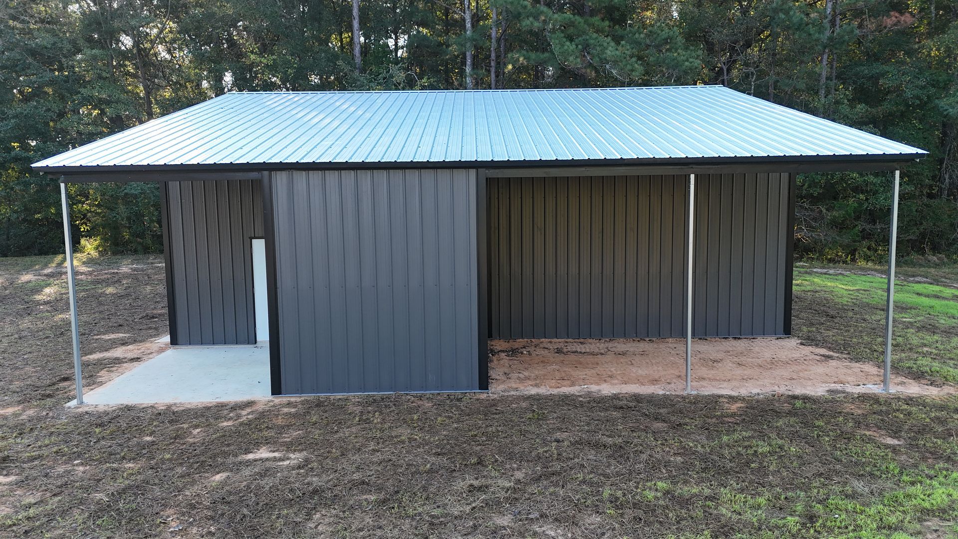 Gray shed with metal roof, open front, set on gravel and concrete pad, surrounded by grass and trees.