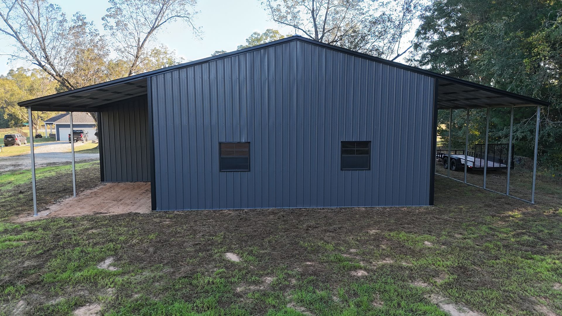 Dark gray metal building with carport, two small windows, gravel base, and green grass.