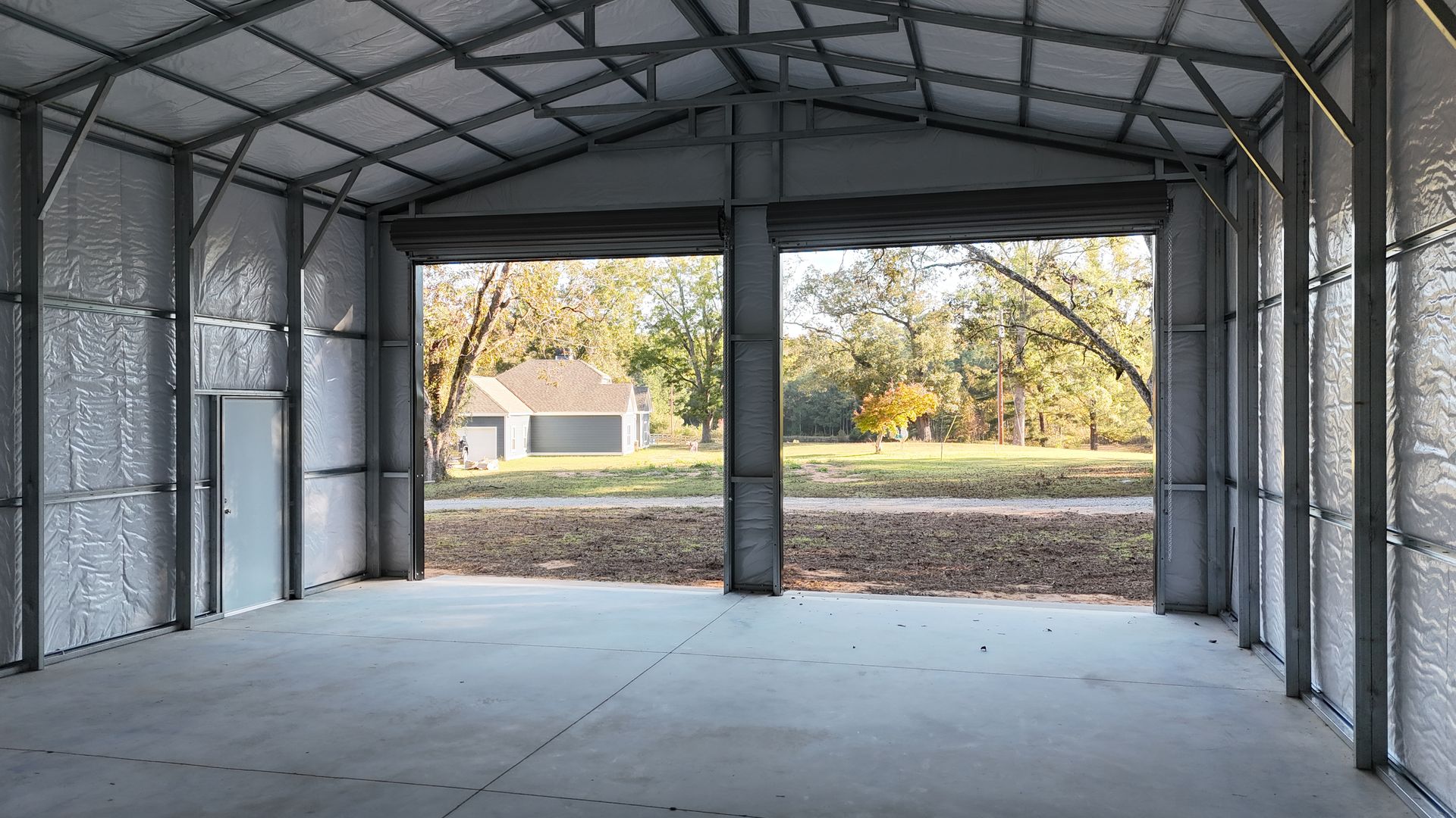 Interior of a metal building with open garage doors looking onto a yard and trees.