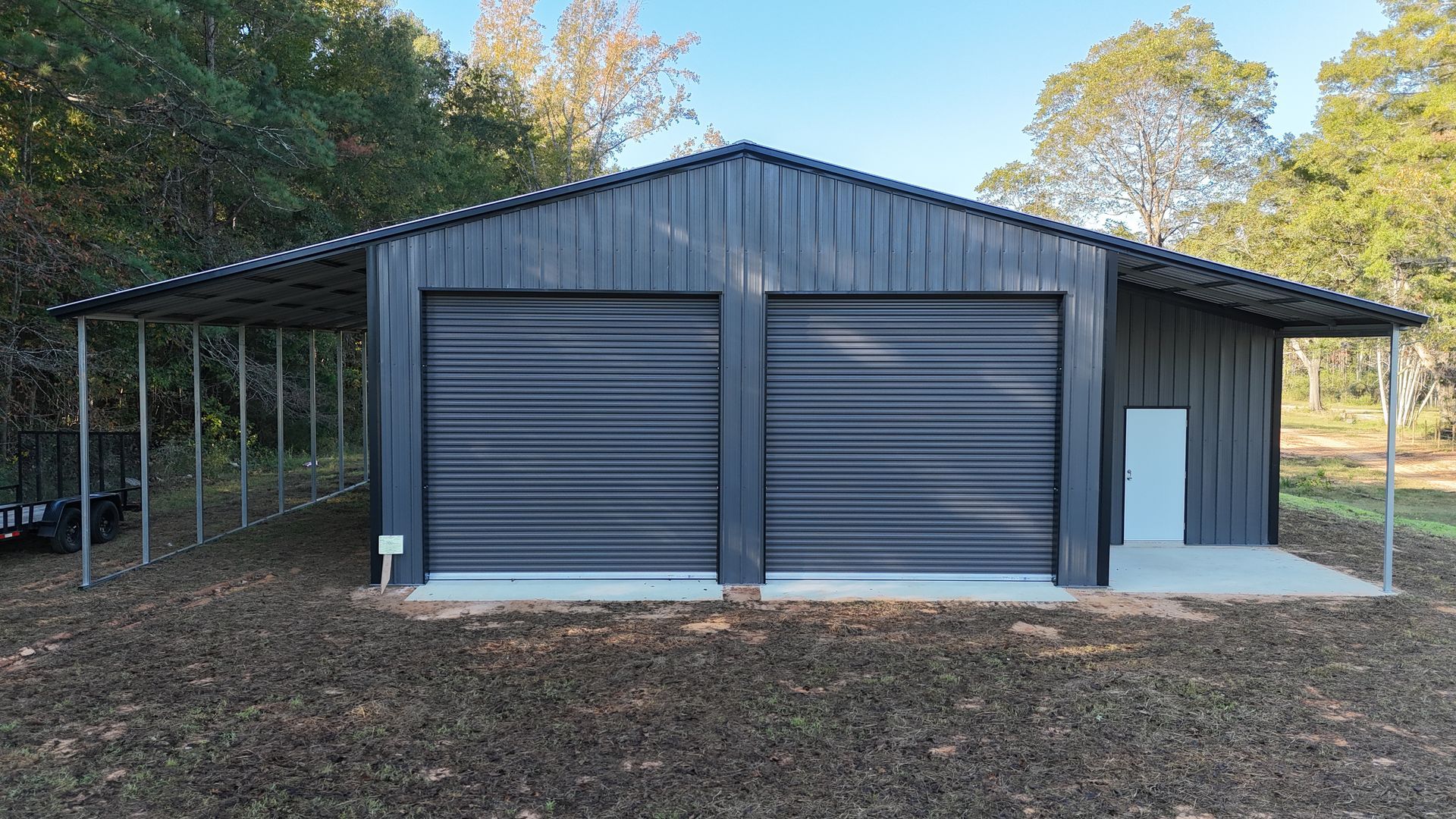 Dark gray metal garage with two doors, a side door, and a covered awning, set in a grassy area with trees.