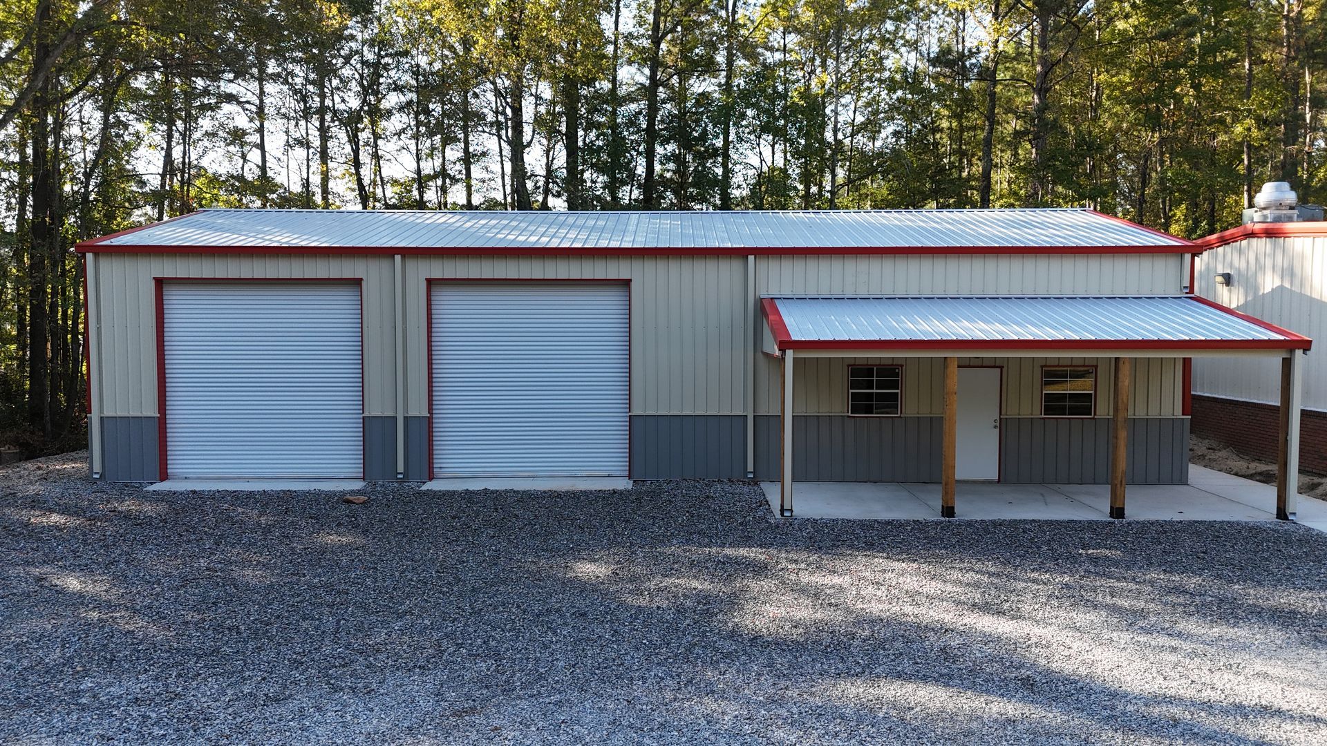 Metal building with two garage doors and a covered porch; gravel driveway, trees in background.
