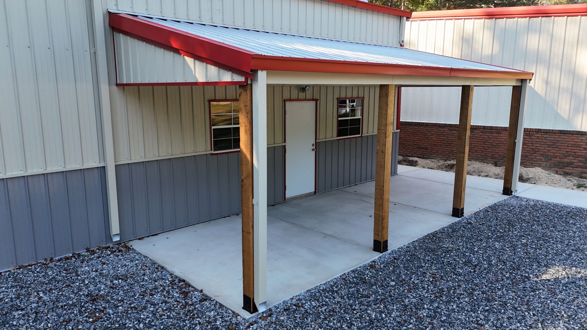 A metal building with a red-trimmed awning supported by wooden posts. Gray and white siding, concrete porch.