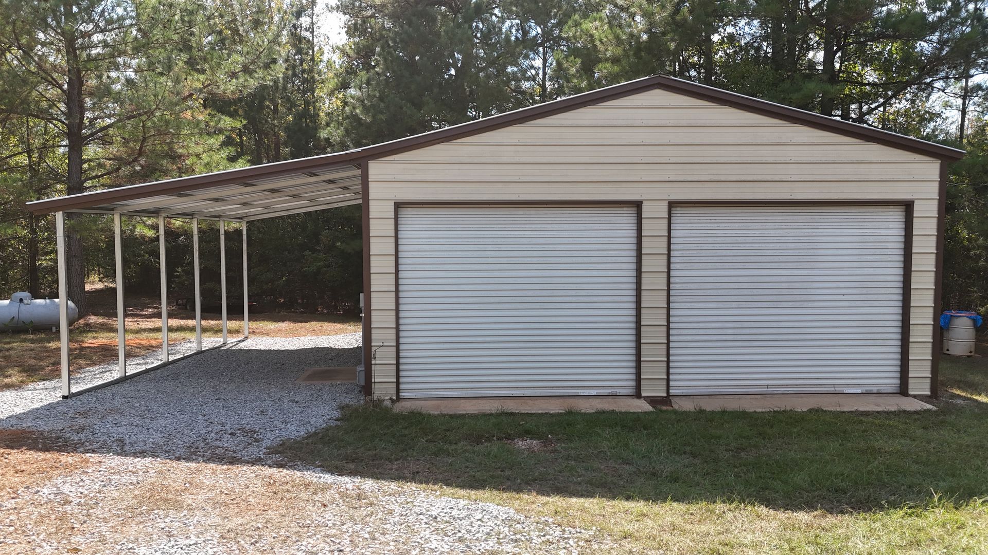 Tan metal garage with attached carport, two roll-up doors, gravel driveway.