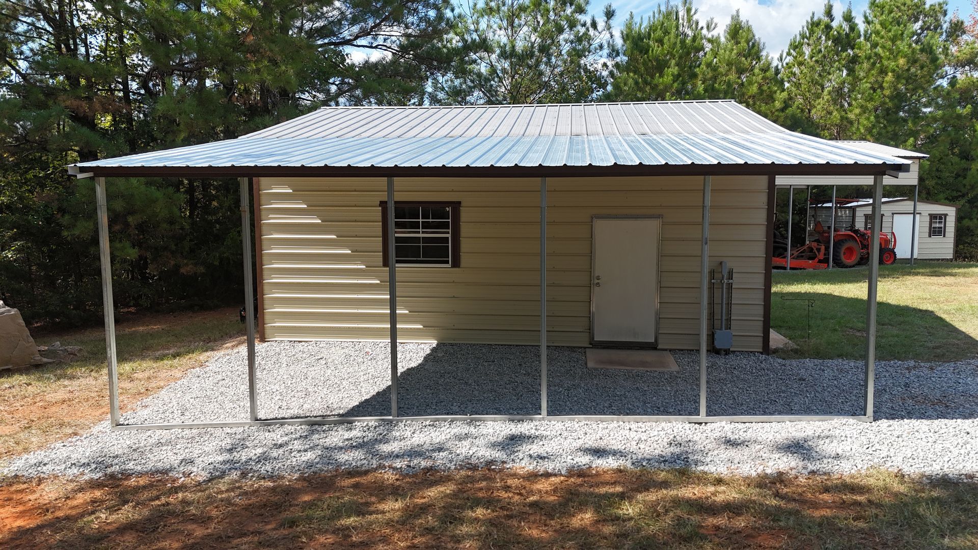 Tan metal shed with silver roof, gravel base, and open front, in a yard with trees.