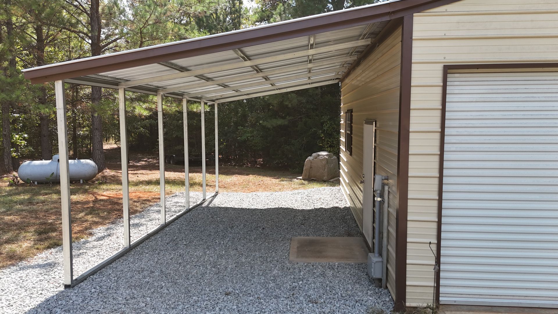 Carport attached to a tan building with a gravel floor, metal roof, and a propane tank.