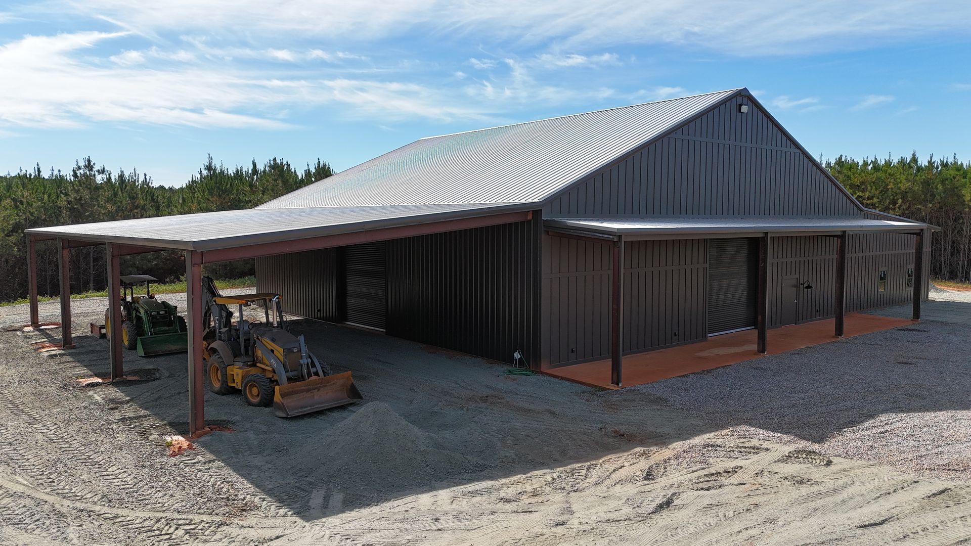 Metal-clad barn with covered porch, gravel driveway, and farm equipment parked outside. Blue sky and trees in the background.
