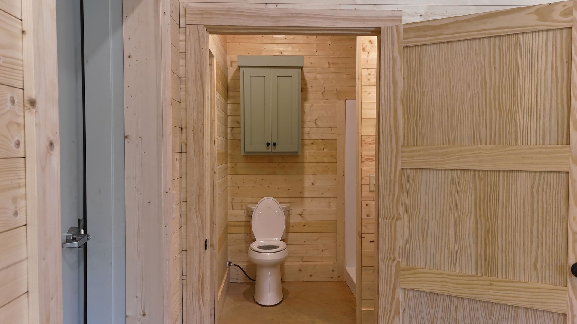 Open doorway to a small bathroom with a toilet, cabinet, and shower. Wooden walls and door.