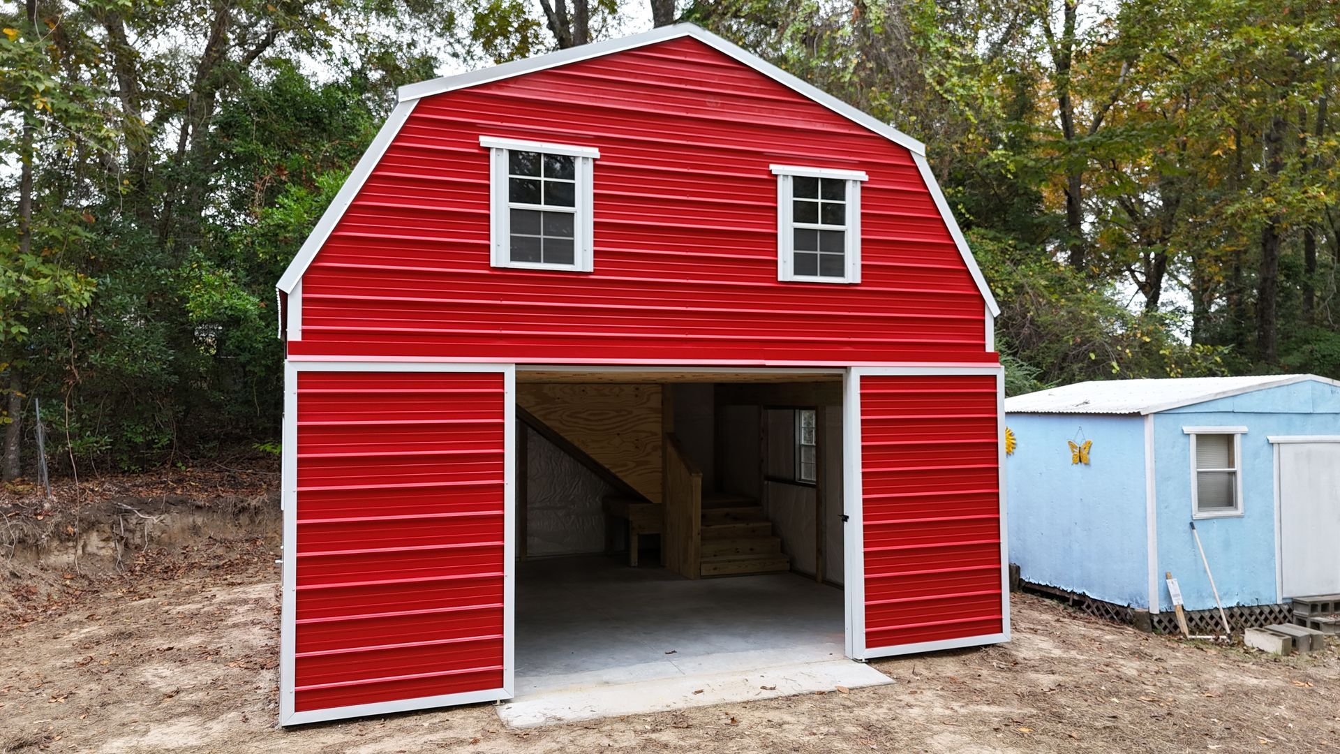 Red barn-style building with white trim and two windows above an open garage. Blue shed next to it.