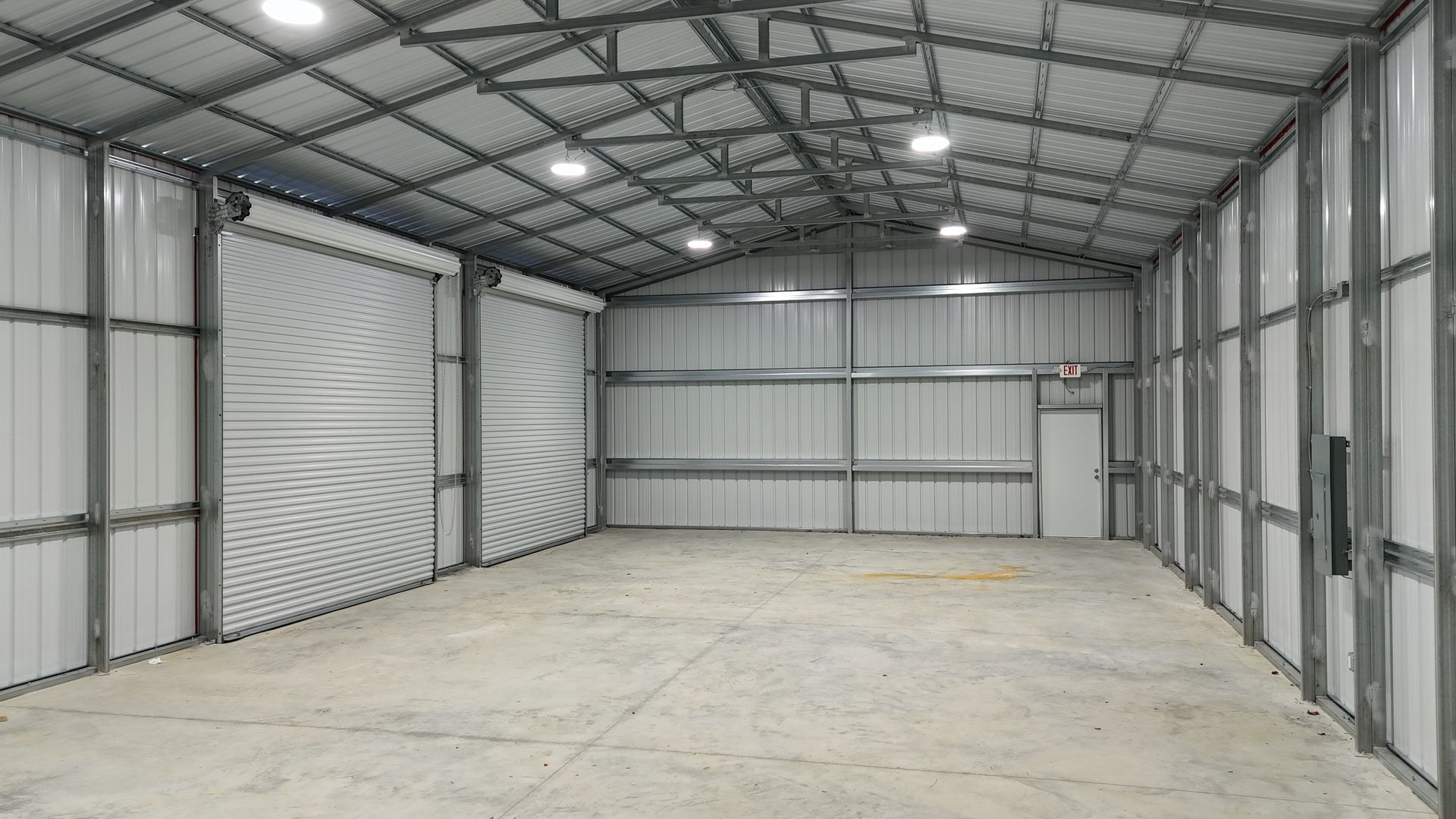 Empty, white-walled metal warehouse interior with a concrete floor, roll-up door, and overhead lights.