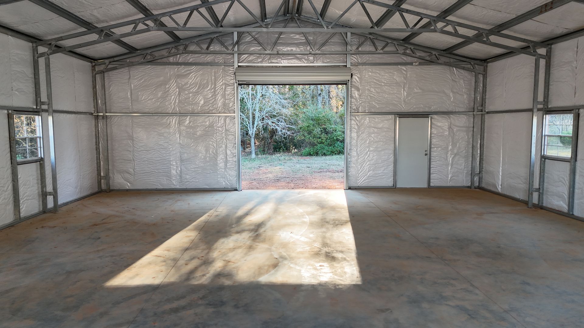 Interior of a metal building with a concrete floor, roll-up door, side windows, and insulation on the walls.