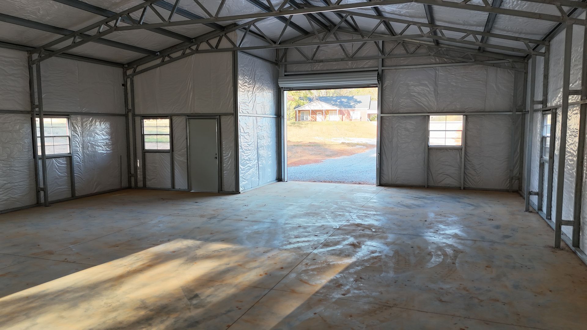 Interior view of a bare metal framed building with concrete floor and roll-up door opening to outside.