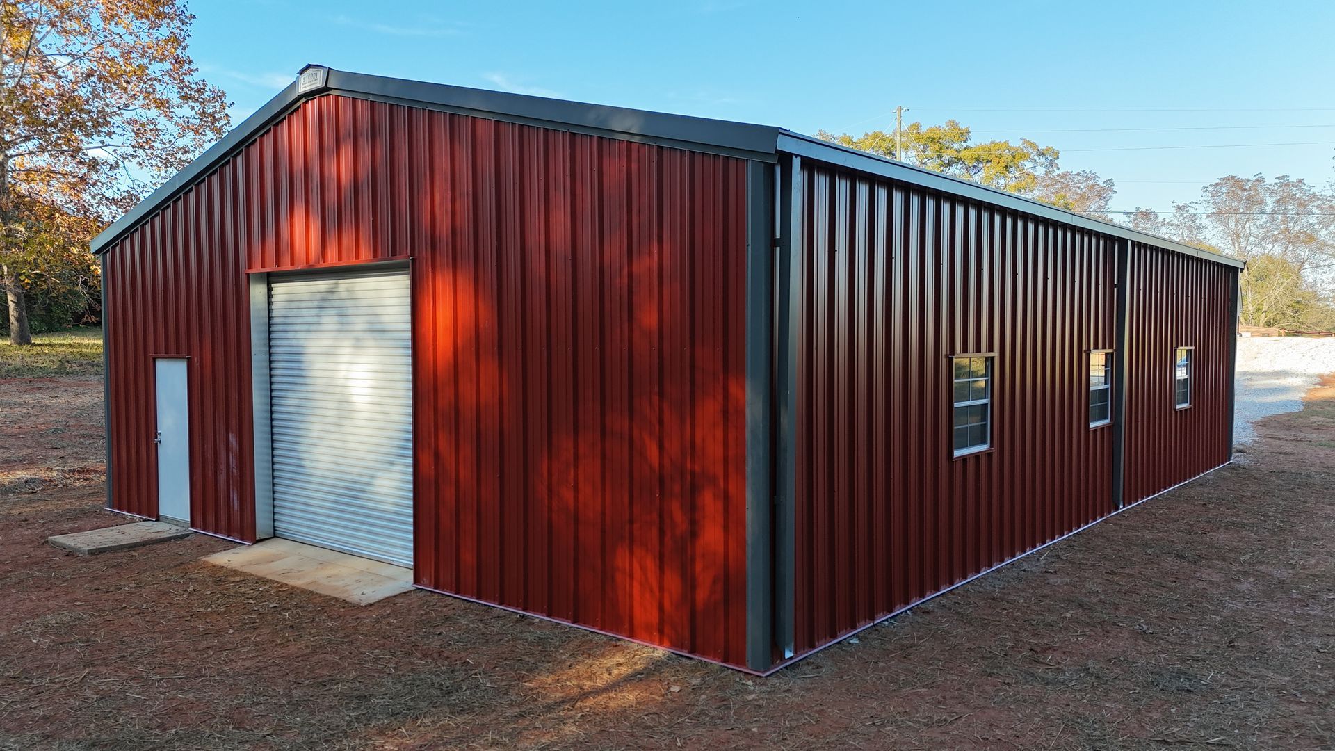 Red metal building with roll-up door, small windows, and dark trim against a blue sky.