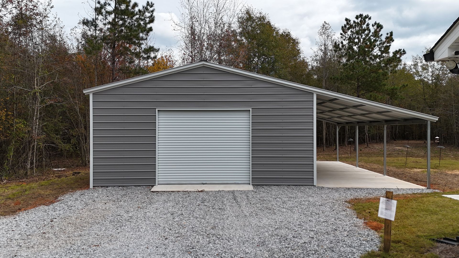 Gray metal garage with attached carport, gravel driveway, and surrounding trees.