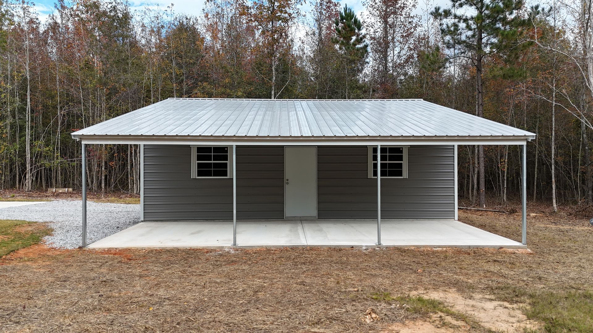 Gray metal building with porch, concrete slab, and trees in the background.