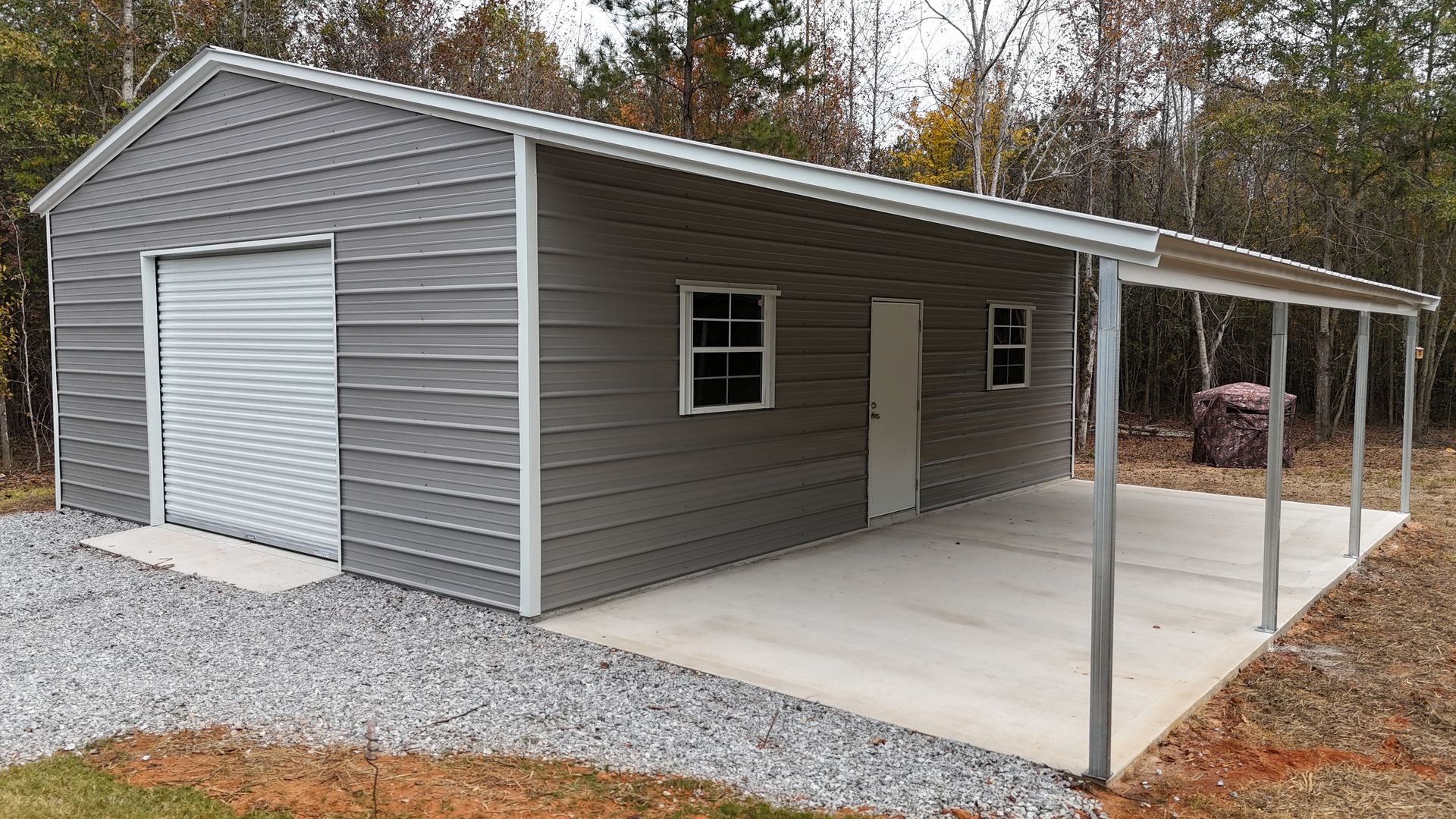 Gray metal building with attached carport; garage door, windows, and concrete pad.