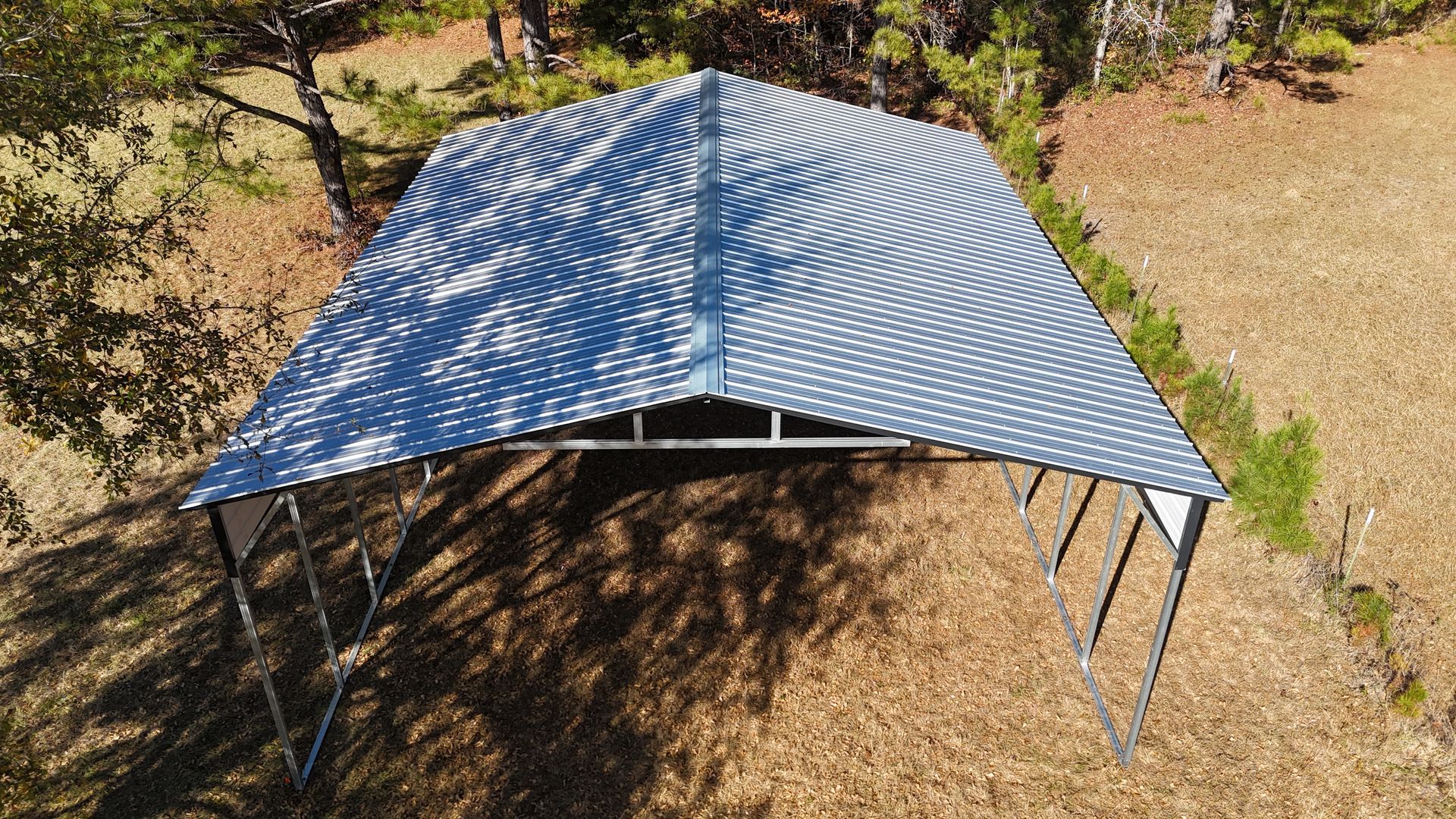 Metal carport in a field, angled roof, blue-gray color, sunlit, surrounding trees and dry grass.