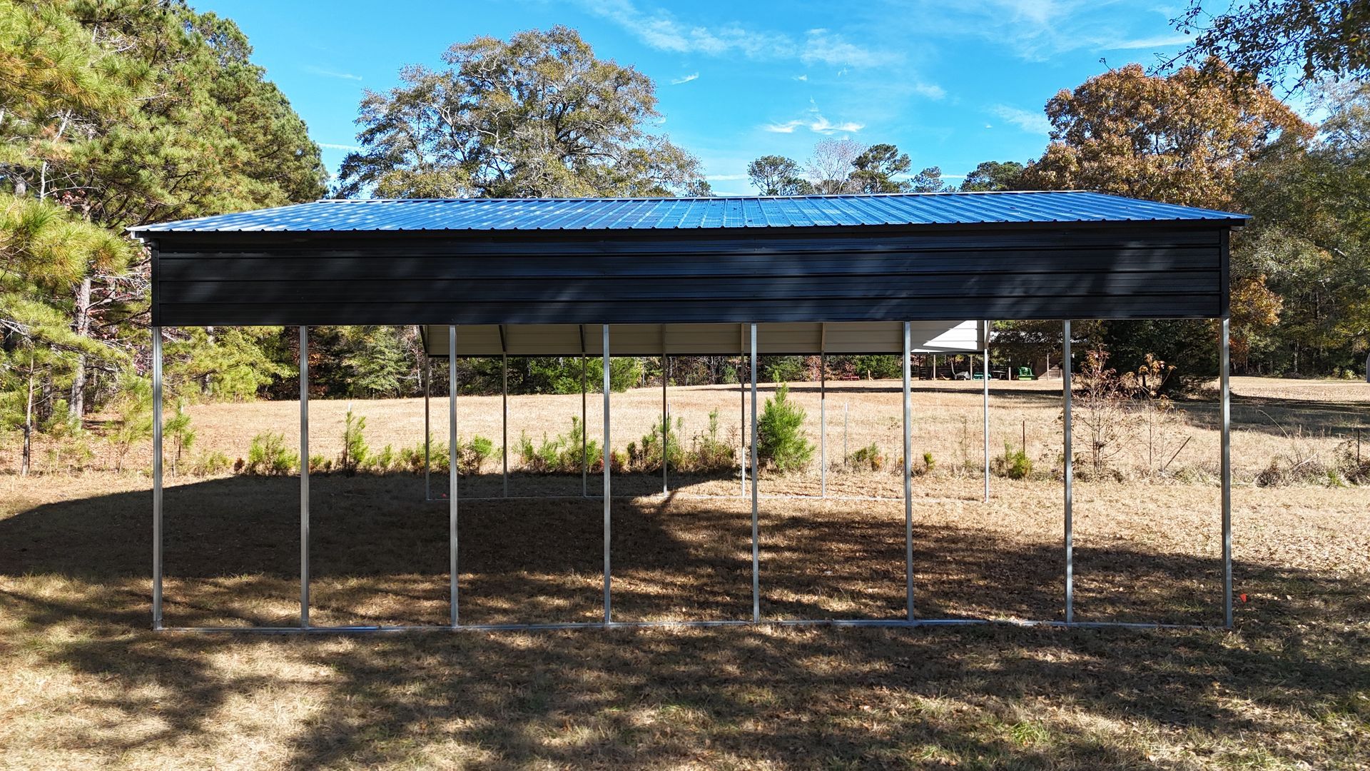 Black carport with blue roof, supported by metal poles, in a grassy field with trees.