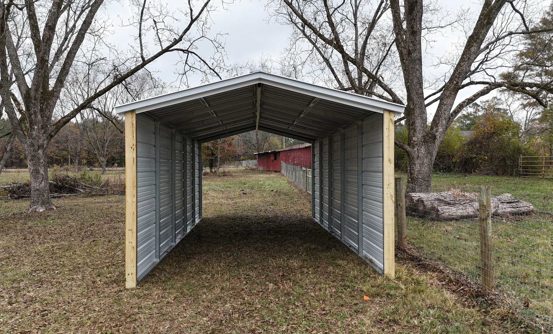 Metal carport on a grassy yard, wooden posts, trees in the background.
