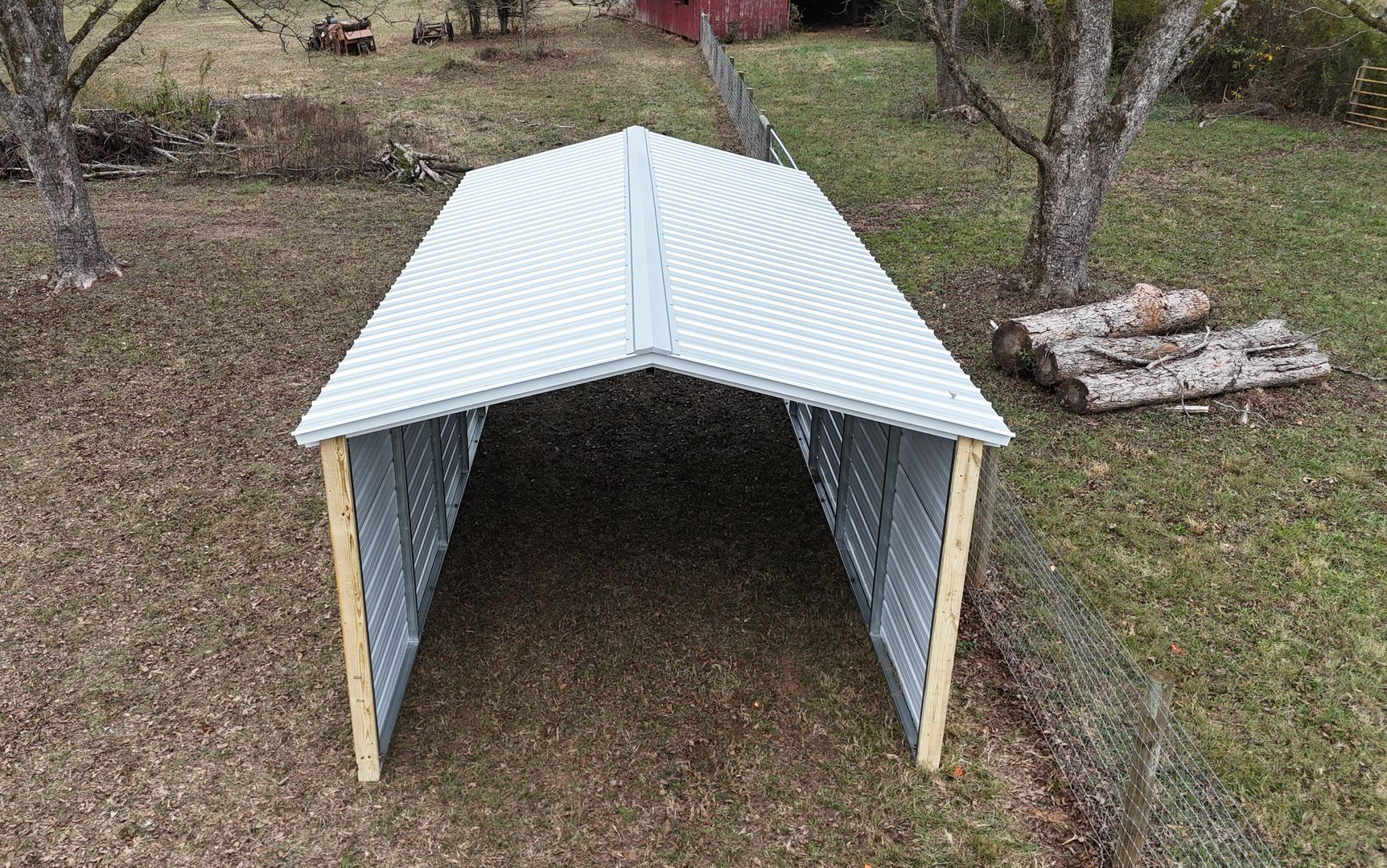 Metal carport with a gray roof and walls, in a grassy area with trees and logs.