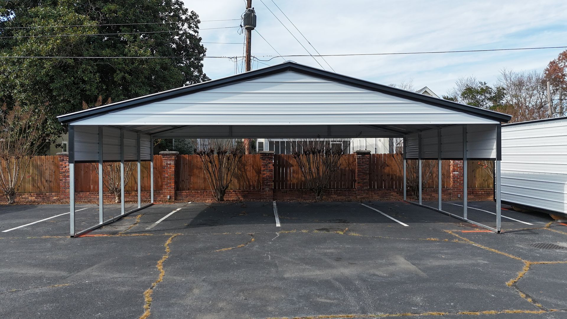 Metal carport over parking spaces, gray roof, white supports, brick fence in background.