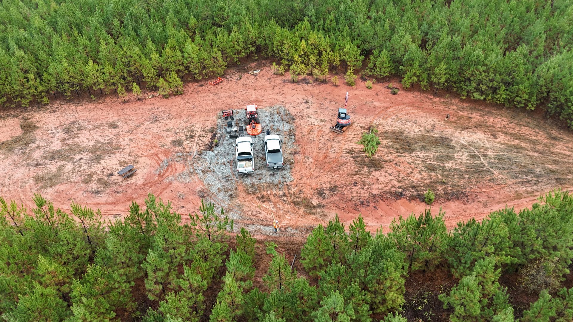 Aerial view of construction site clearing a red-brown dirt path in a green forest. Trucks and equipment are present.