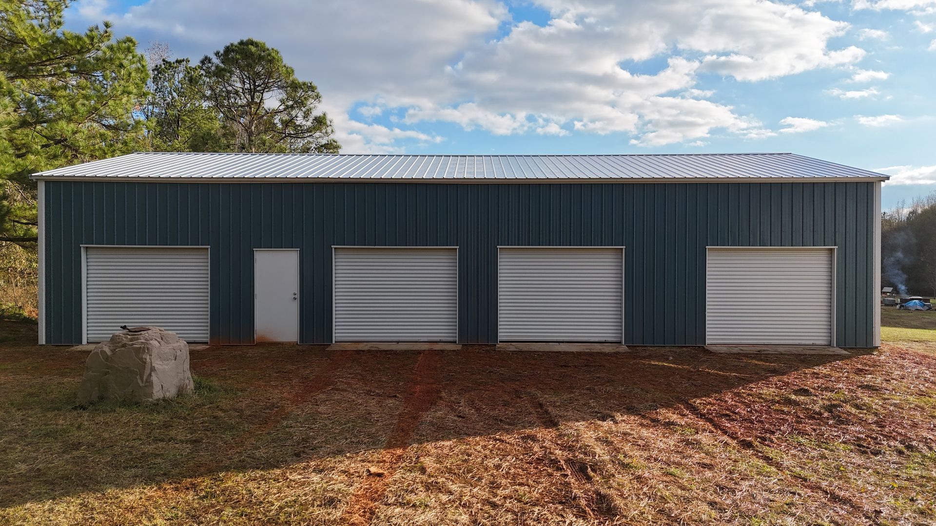 Blue metal building with four garage doors and a white door, in front of trees.
