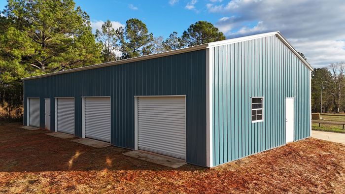 Blue metal storage building with four roll-up garage doors, a side entrance, and a window, situated on a wooded lot.