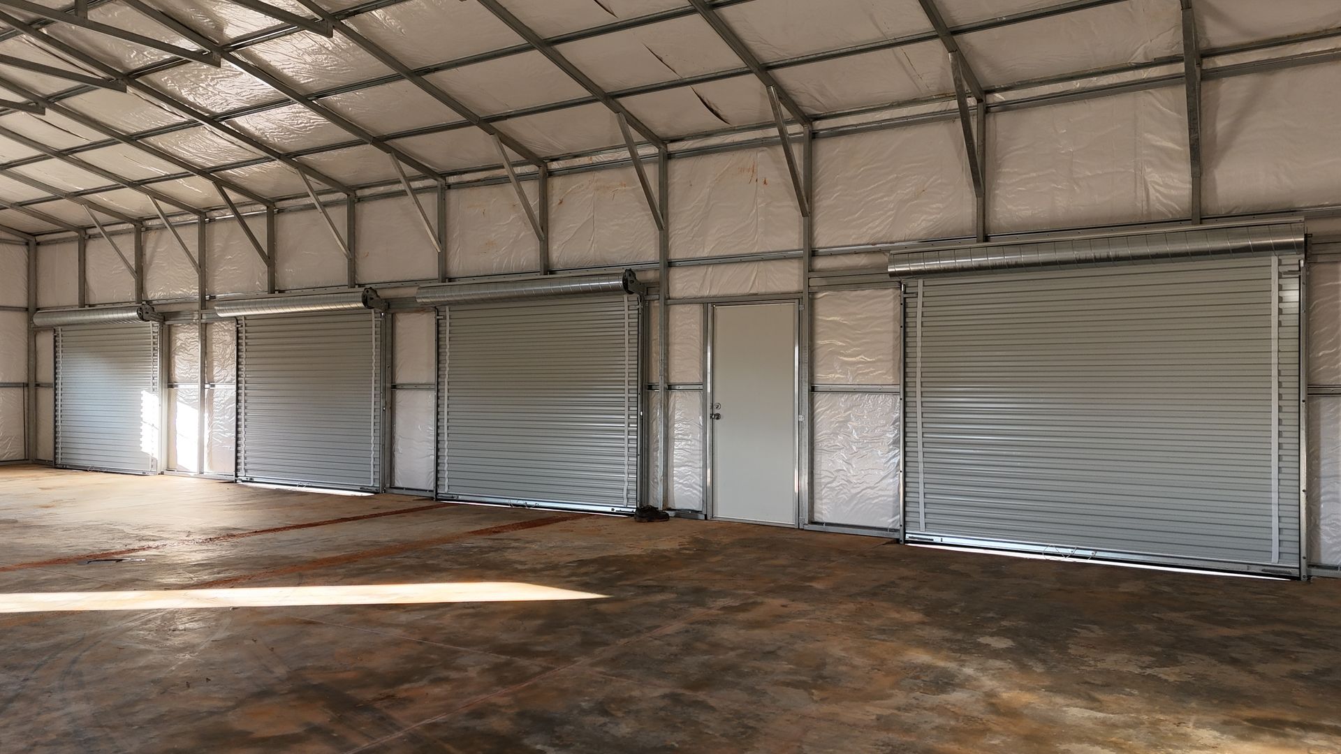 Interior of a metal building with three roll-up doors and a side door, illuminated by sunlight.