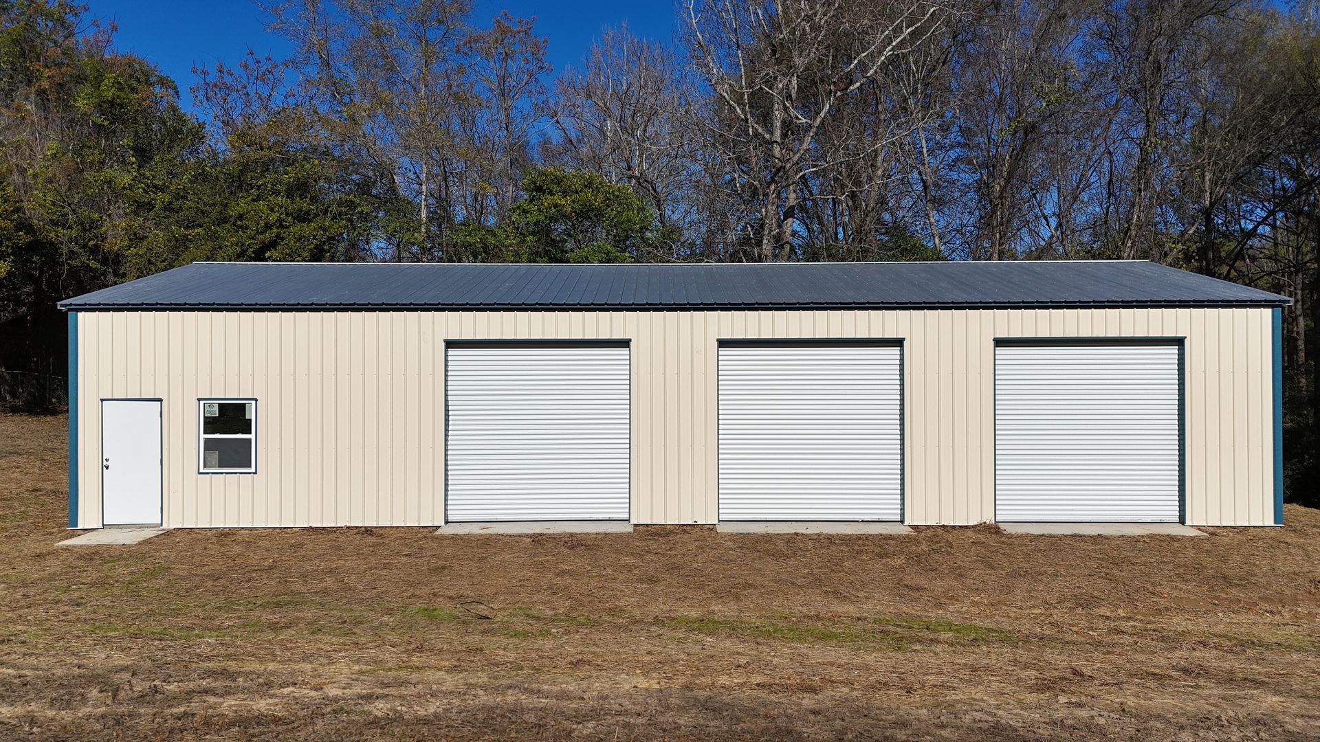 Tan metal garage with three bays and a single door, set in a field with trees in the background.