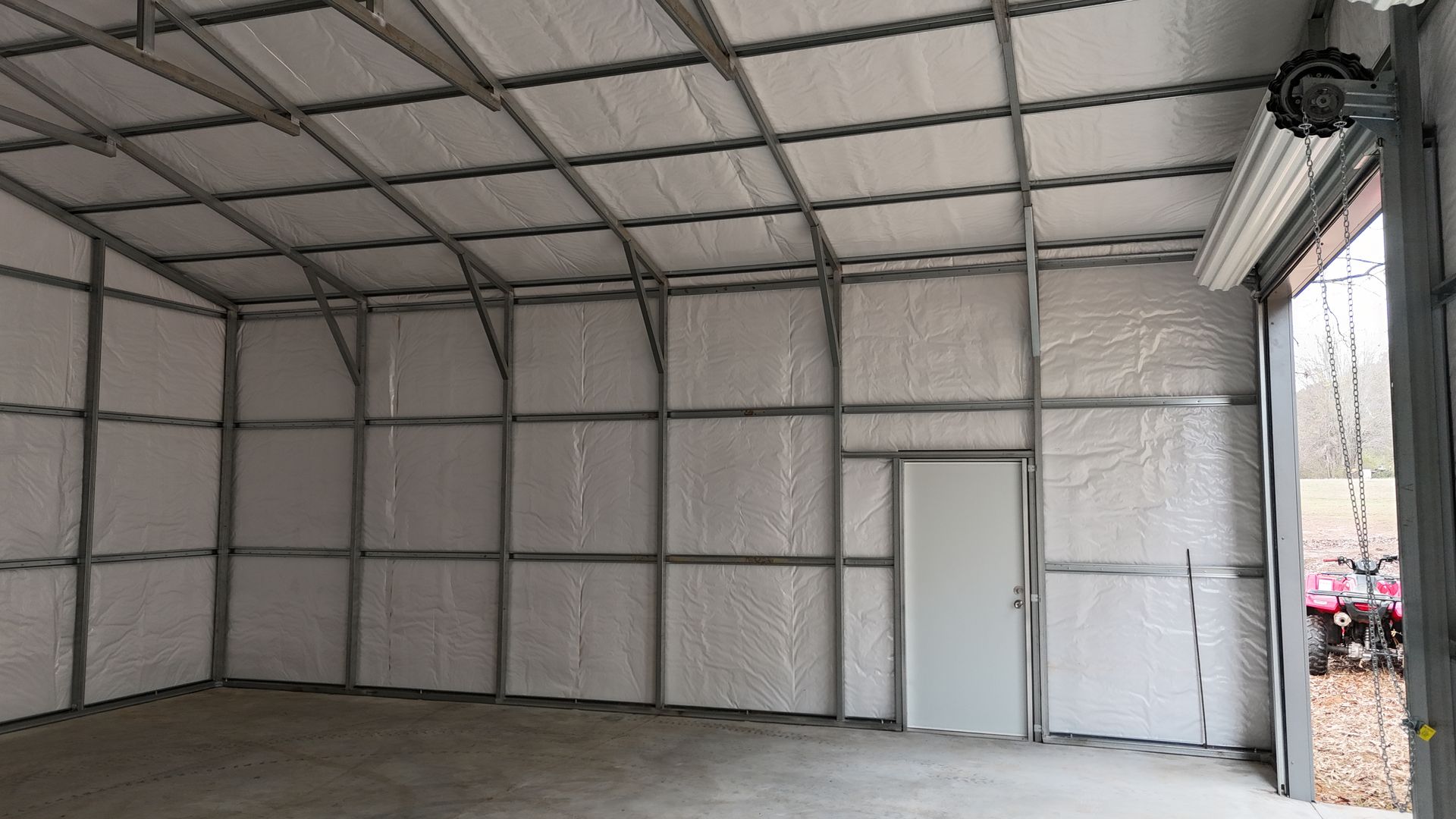 Interior view of a metal building with insulation, a closed door, and an open garage door. Concrete floor.