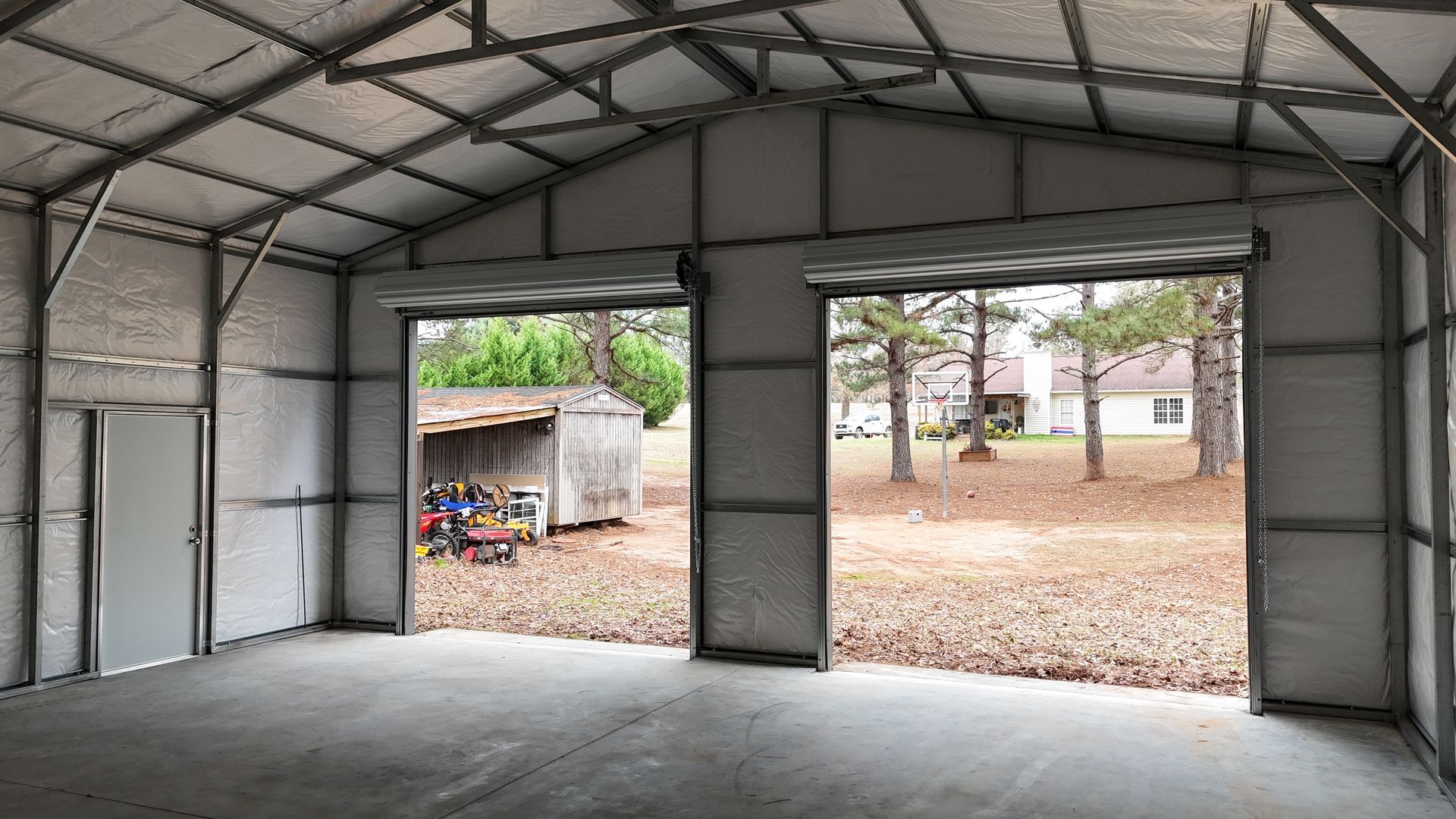 Interior of metal building with open roll-up doors, concrete floor, and small shed visible outside.