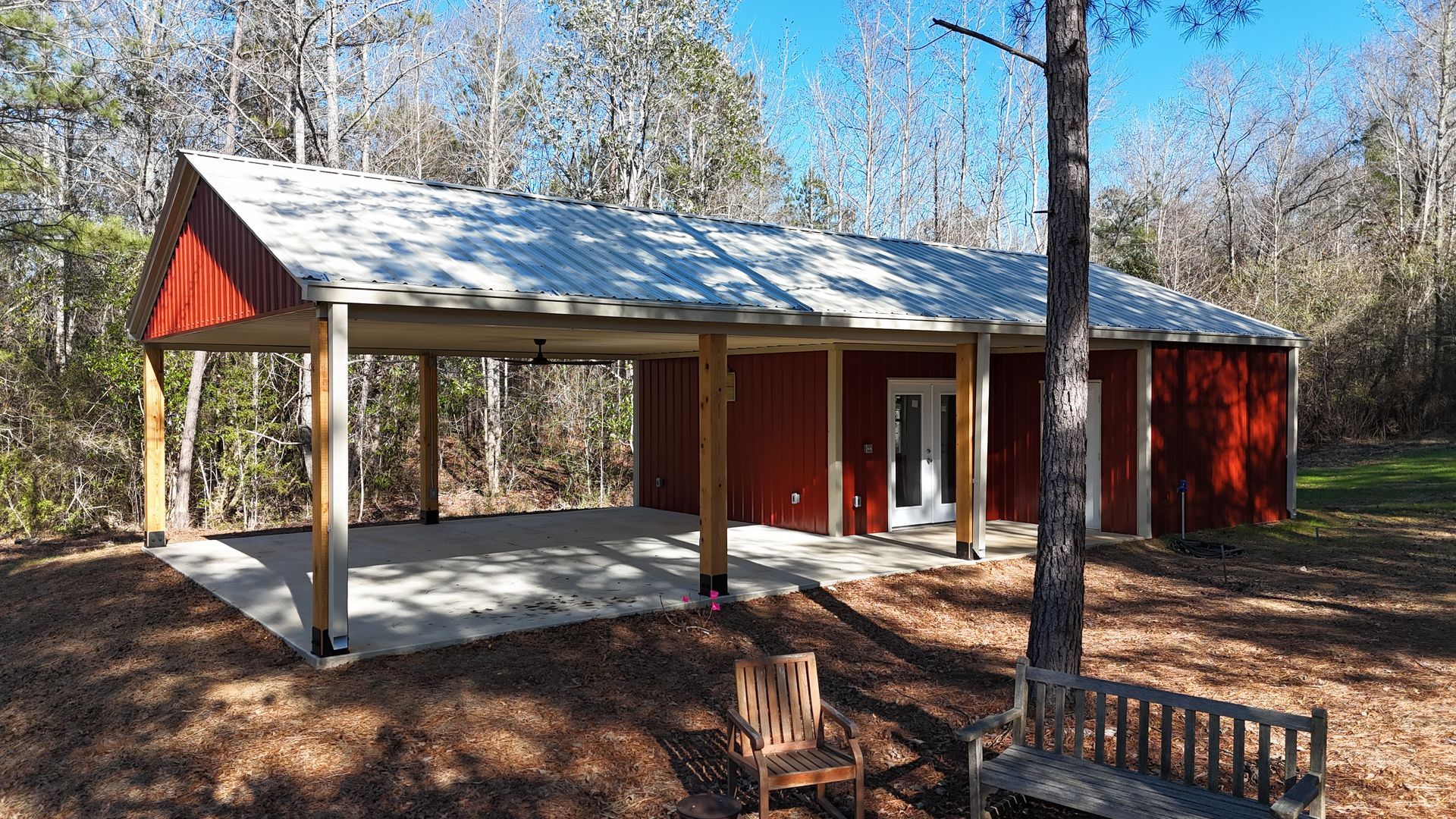 Red building with carport, metal roof, concrete patio, in a wooded setting.