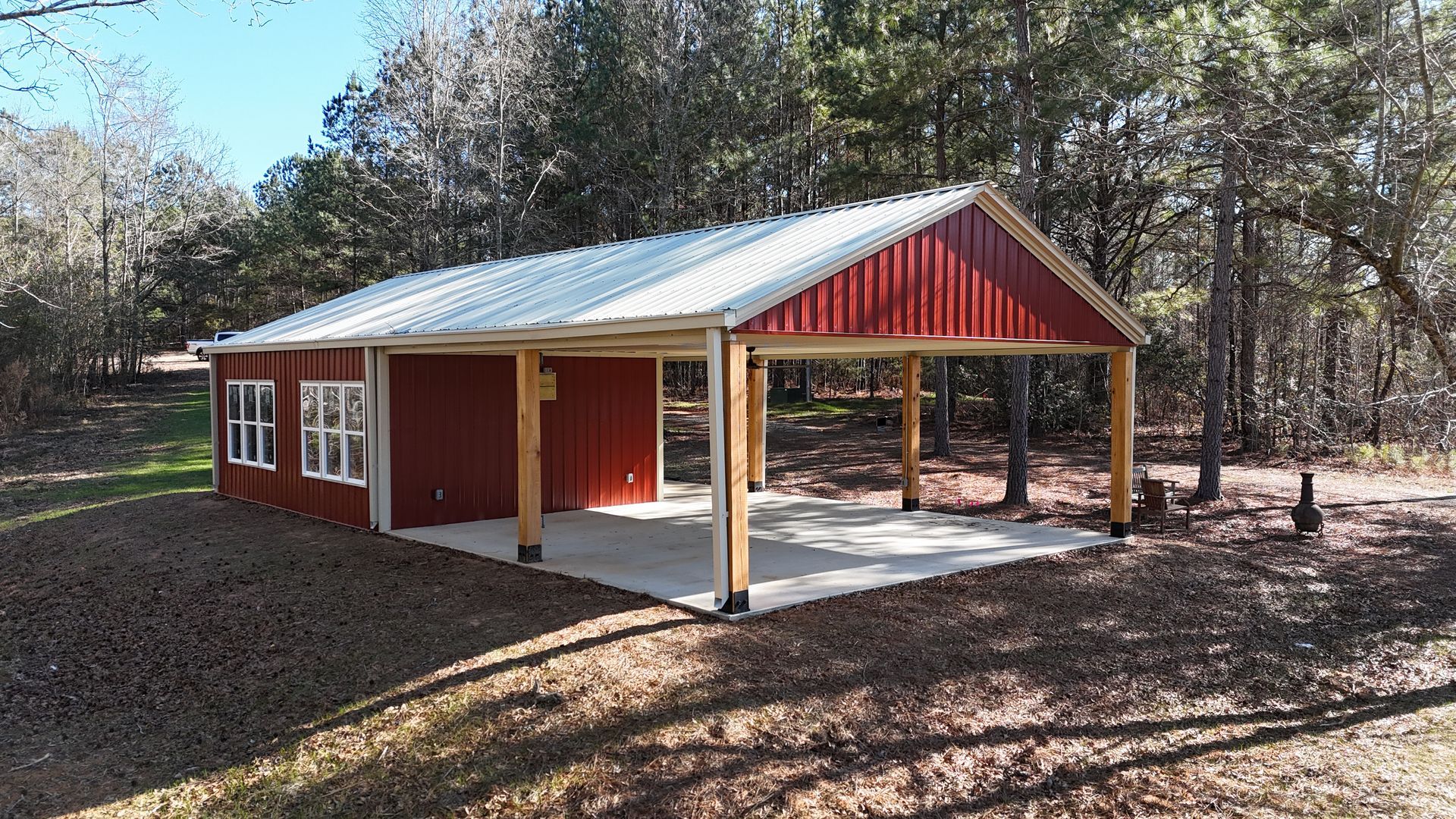 Red and white shed with a carport, set in a wooded area.