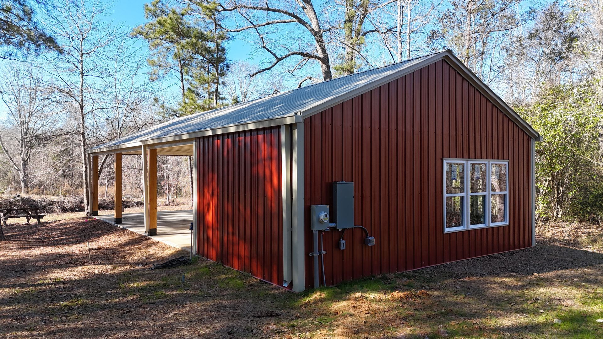 Red metal-sided building with a covered porch in a wooded setting under a blue sky.