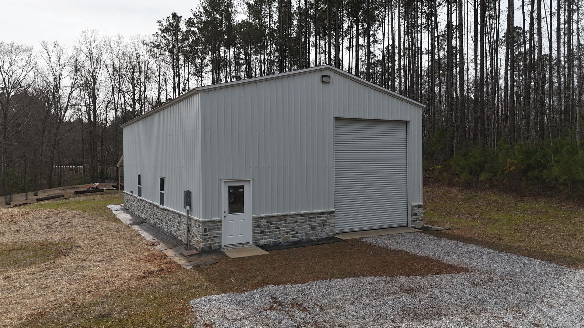 Metal building with stone base, large roll-up door, small entry door, gravel driveway, surrounded by trees.