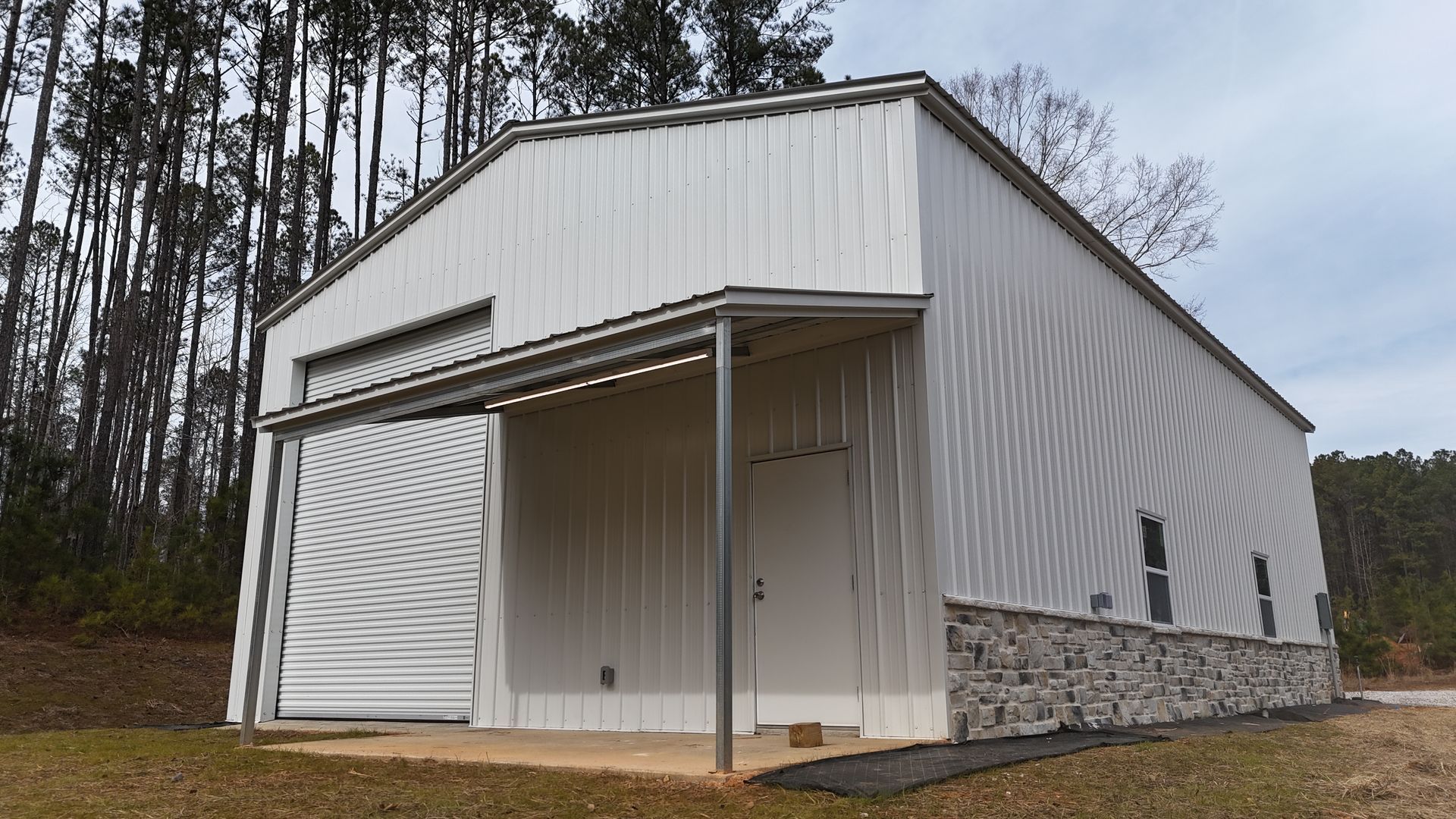 White metal building with a covered porch and stone base, set in a grassy area with trees in the background.