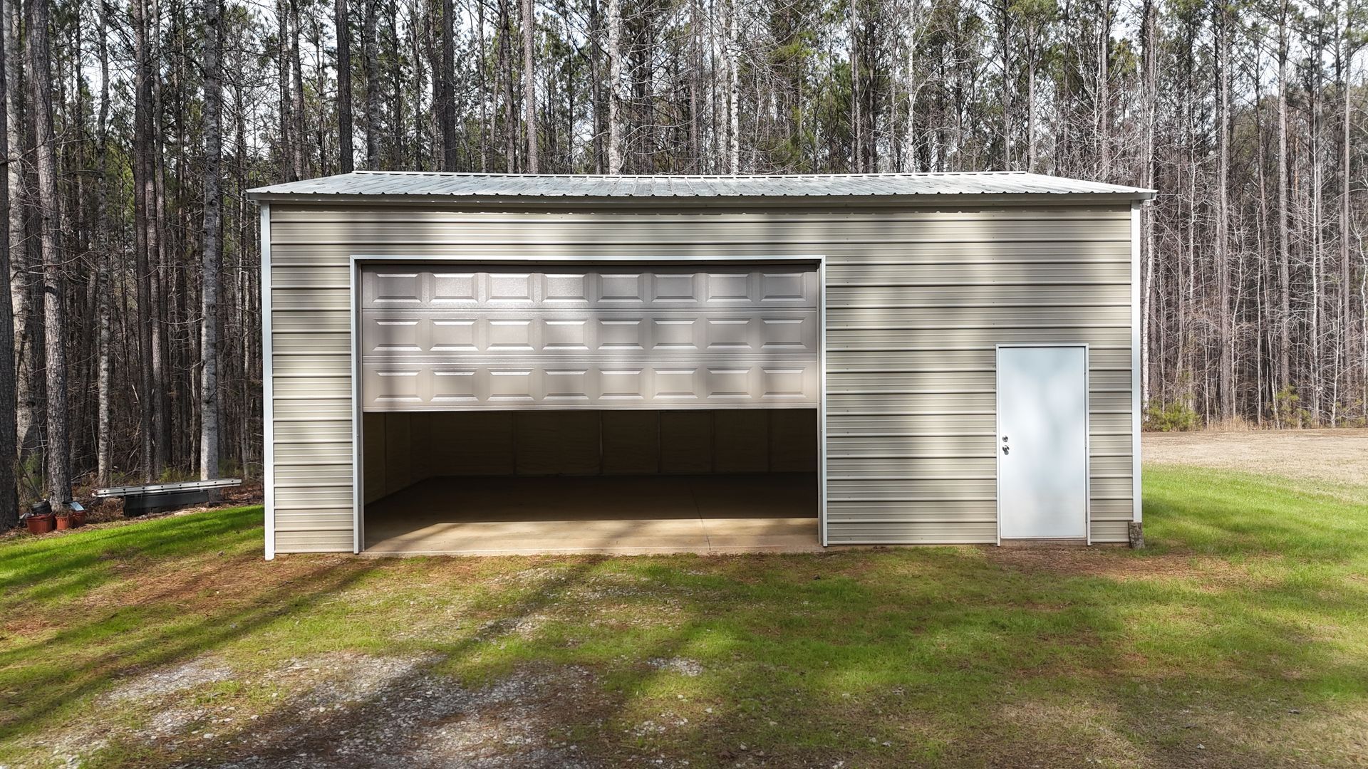 Gray metal garage with open door, small side door, and corrugated metal roof. Located in a grassy area with trees.