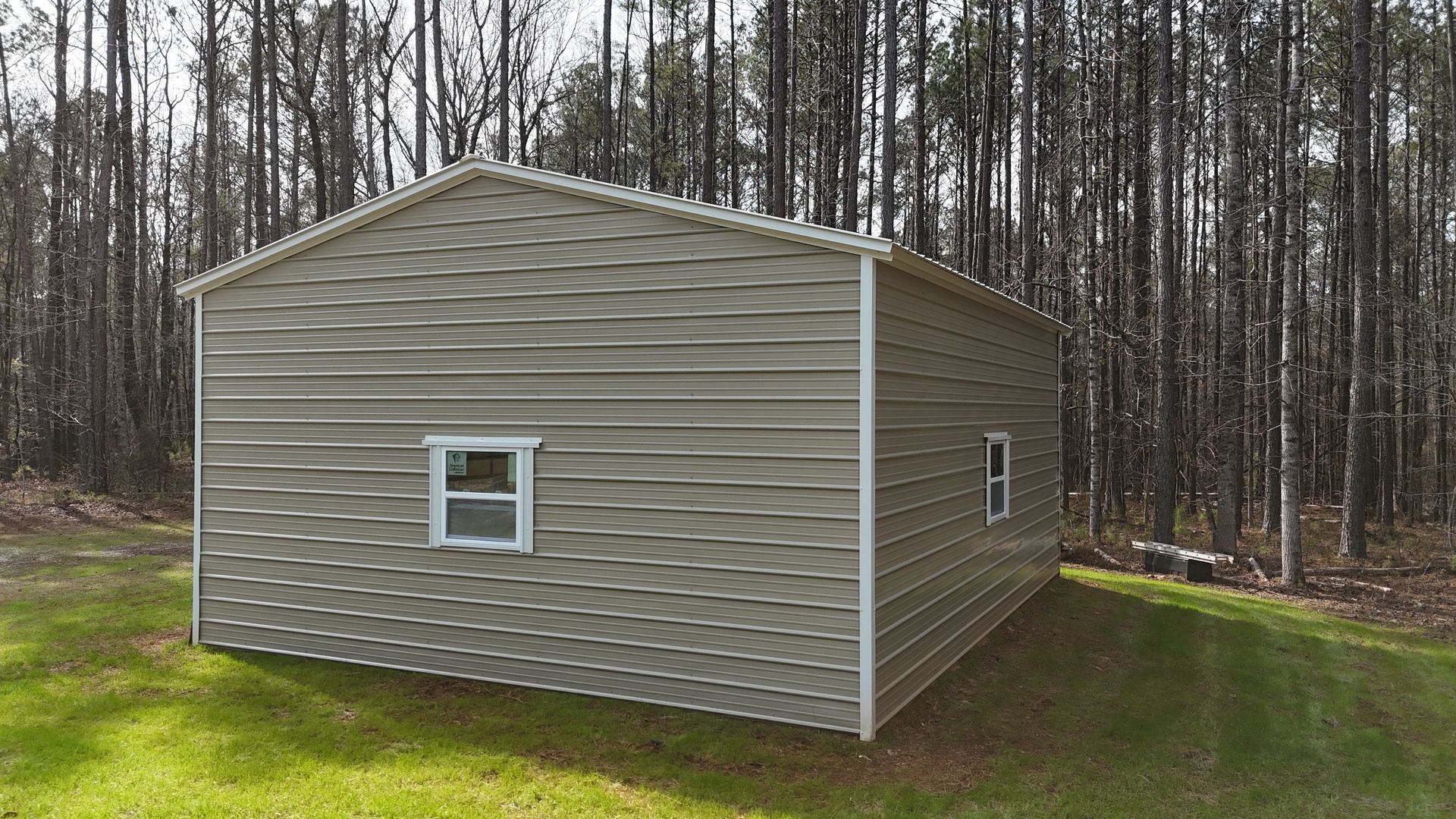 Tan metal shed with two windows, on green grass, wooded background.